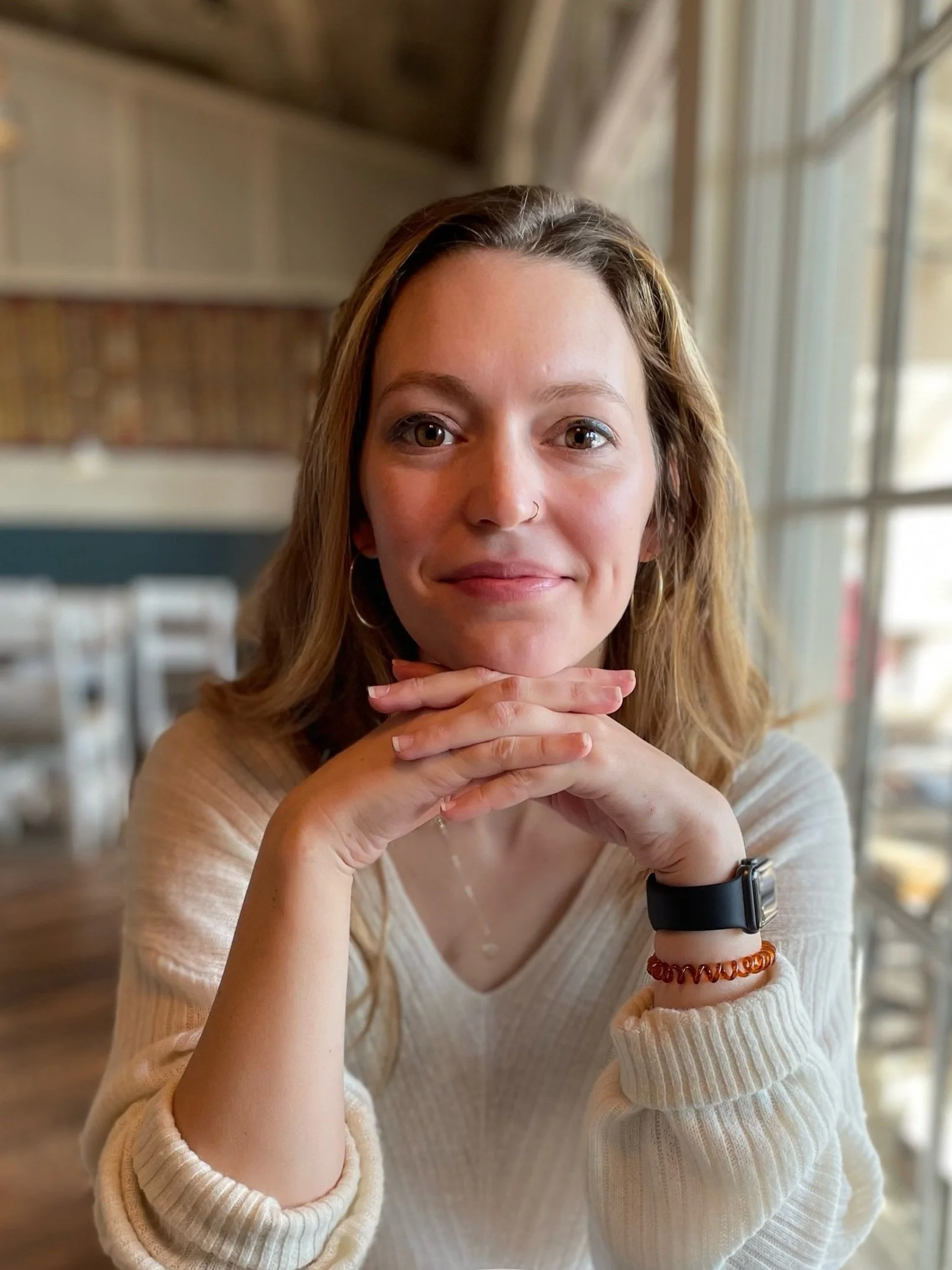 A person with light brown hair, wearing a white sweater, sits indoors with hands clasped under their chin, smiling at the camera. The person has a nose ring and is wearing a black smartwatch and an amber bracelet. The background shows a window and blurred interior.
