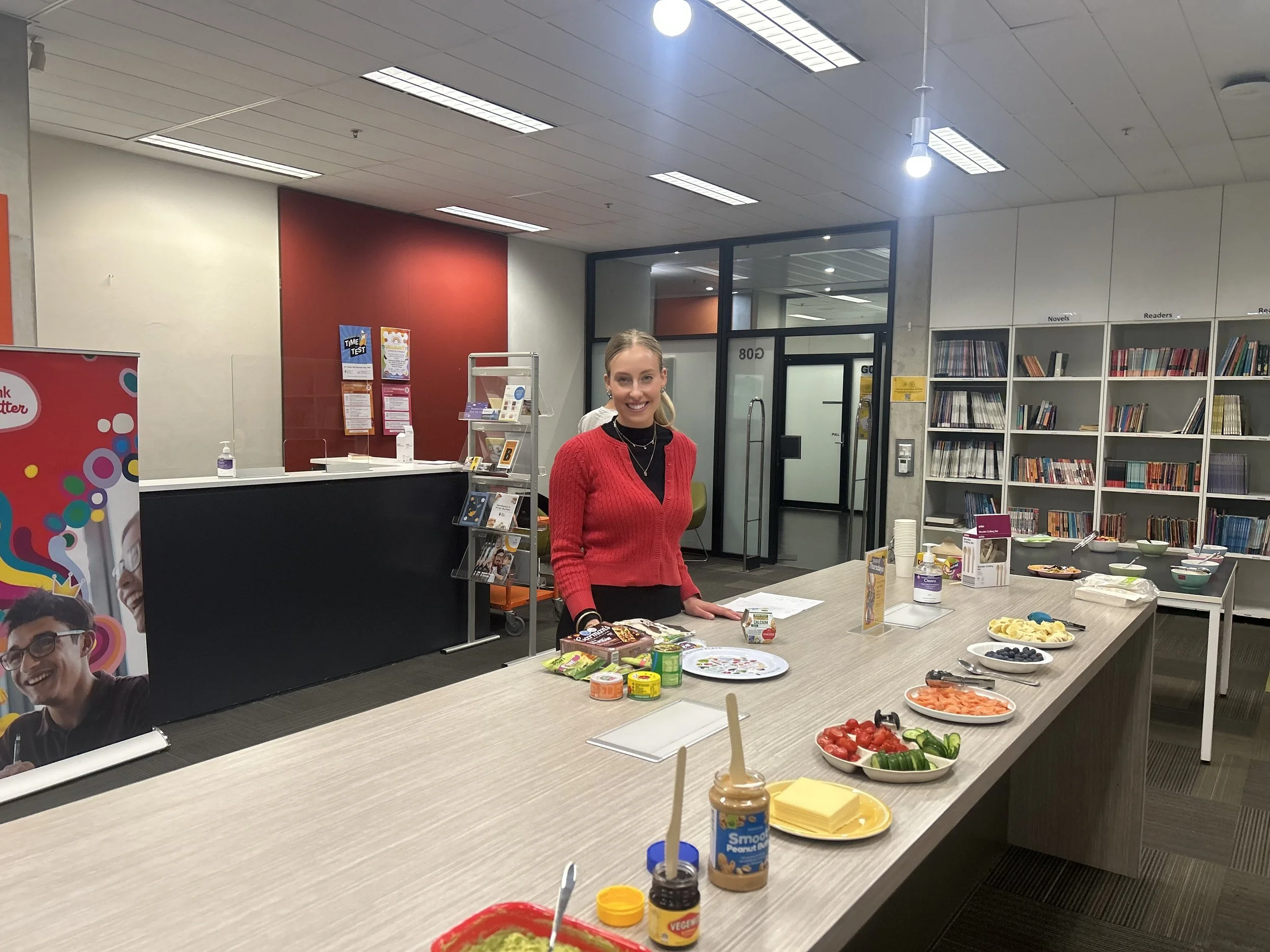 A woman in a red sweater standing behind a table with various food items in a library or bookstore setting