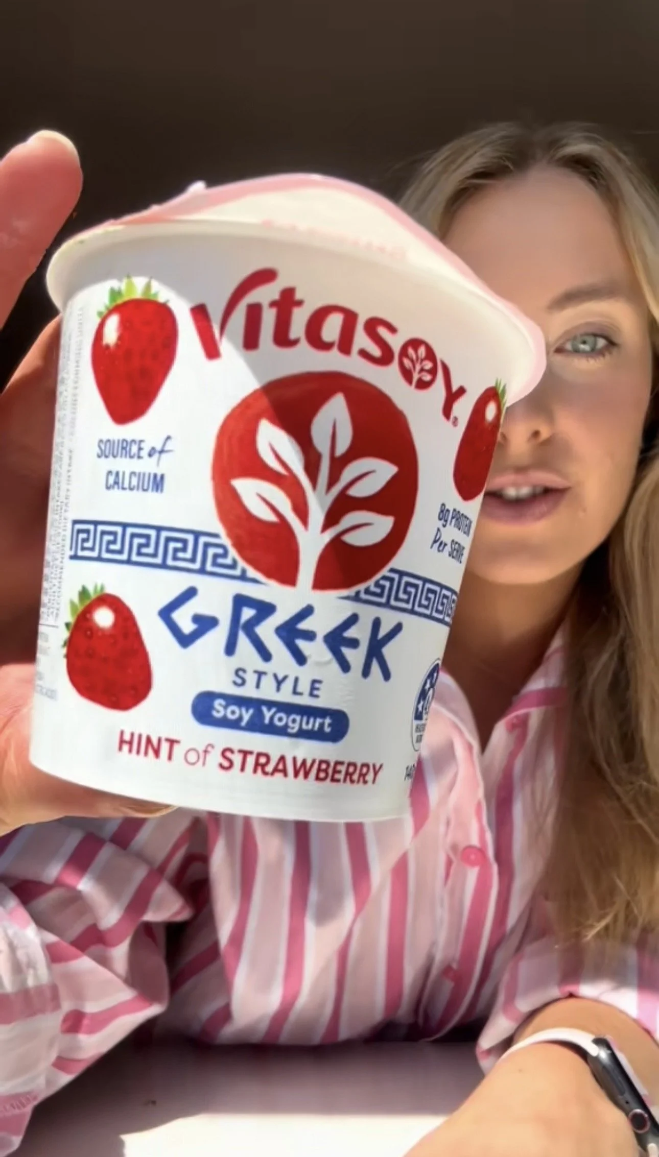 A woman holding a container of Vitasoy Greek Style Soy Yogurt with a hint of strawberry, partially obscuring her face. She is wearing a pink and white striped shirt.