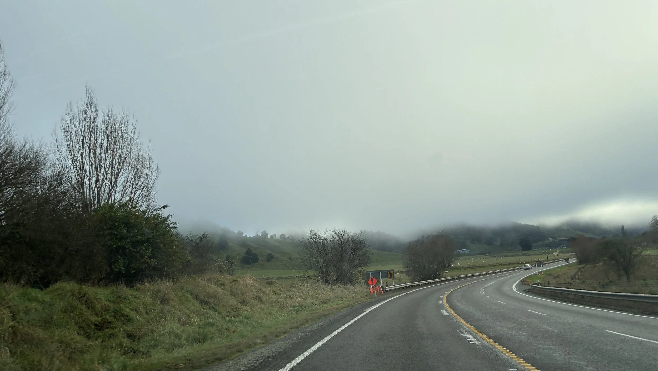 A road turning to the right, with farmland and trees on either side. The top half of the scene is covered in grey low cloud.