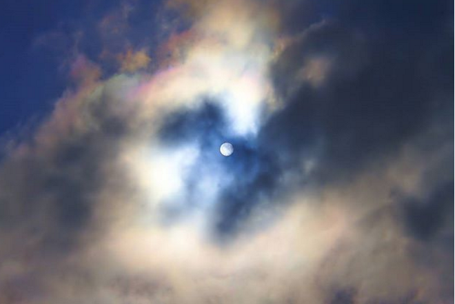 The moon shining through wispy clouds. Rainbow patterns appear in the clouds.