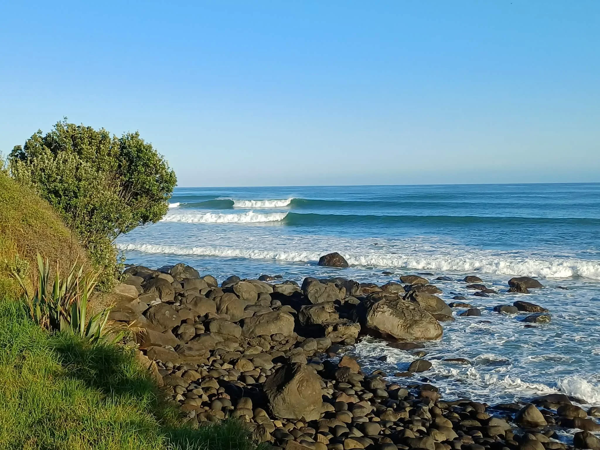 A seaside scene, with cloudless blue sky. The beach is rocky, with a grassy hill on the left side. The sea is very blue and has two distinct breaking waves, with a third frothy line at the edge of the rocks.