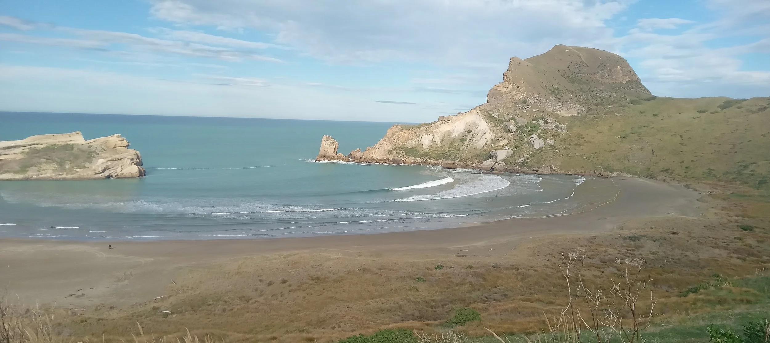 A seaside scene, with a sandy beach. The sea spills between two rocky arms into a more sheltered cove. A person is barely visible in the distance, in the bottom-left corner of the image.