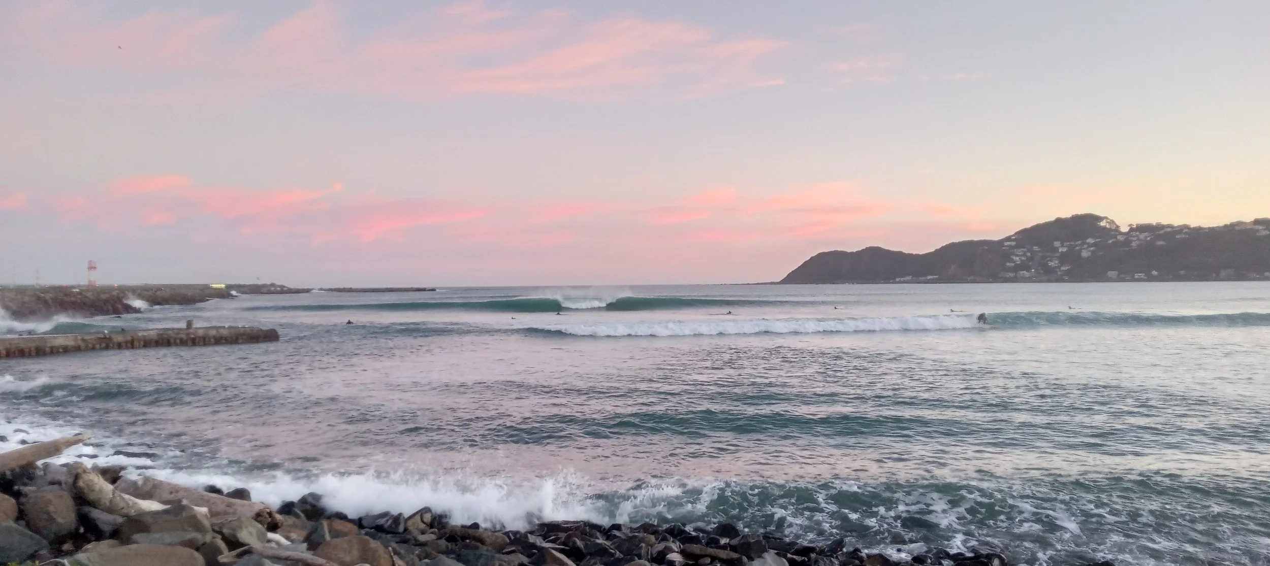 A seaside scene, framed on the left by a long, flat promontory, and on the right by distant hills. Two distinct breaking waves are visible. A number of surfers are waiting to catch one wave, while one is surfing the other wave.