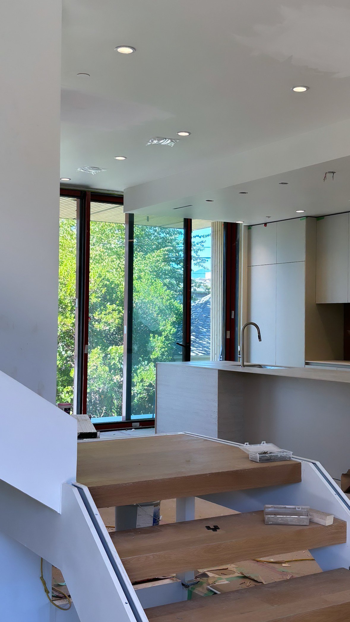 Modern interior with unfinished wooden stairs and large windows, overlooking a green view, featuring minimalist kitchen with white cabinetry and island.