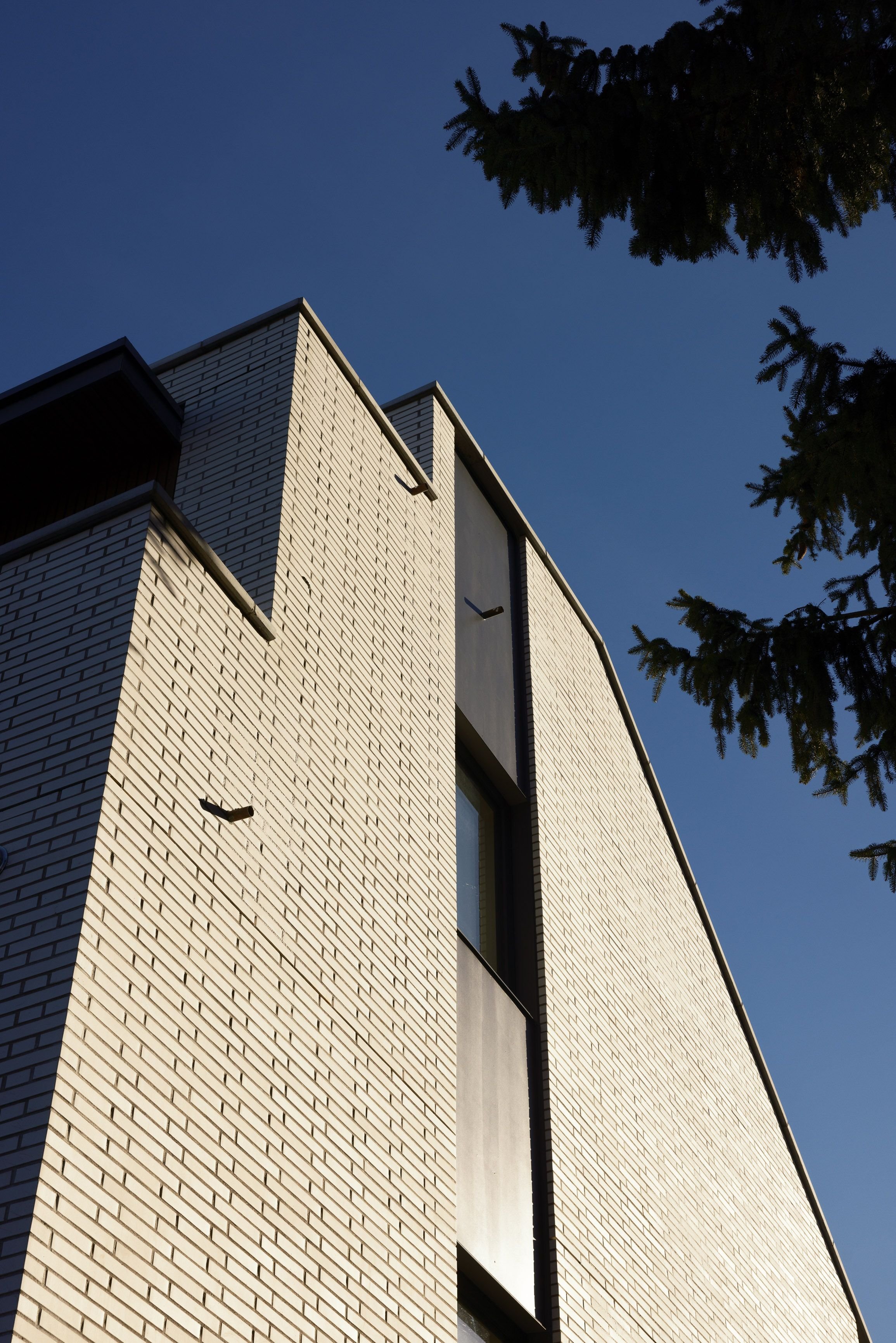 Angular view of a modern brick building with a clear blue sky and tree branches in the foreground. The building features a large window and minimalist design.