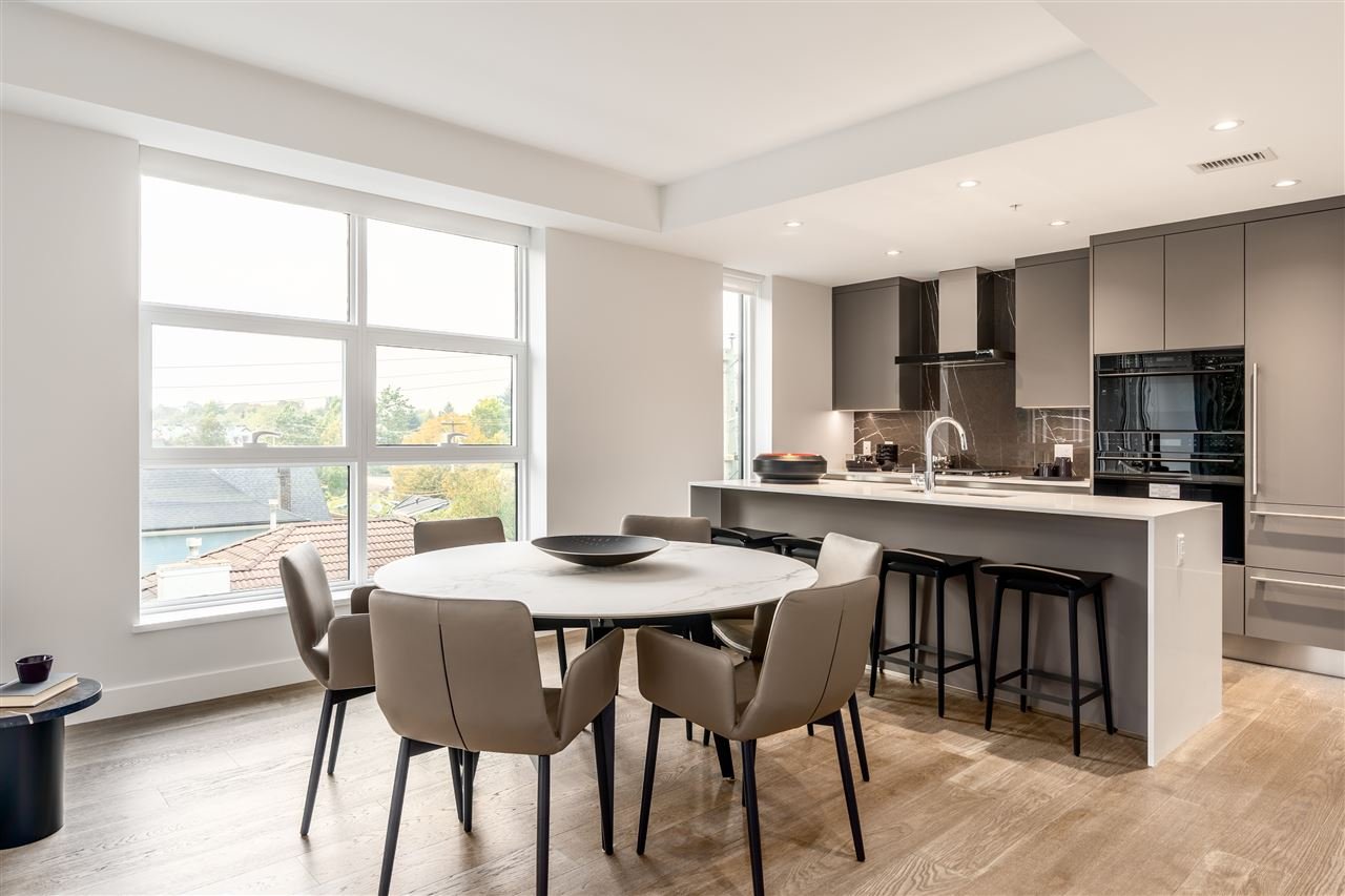 Modern kitchen and dining area with a round table, beige chairs, and a breakfast bar with stools, featuring neutral tones and large windows.