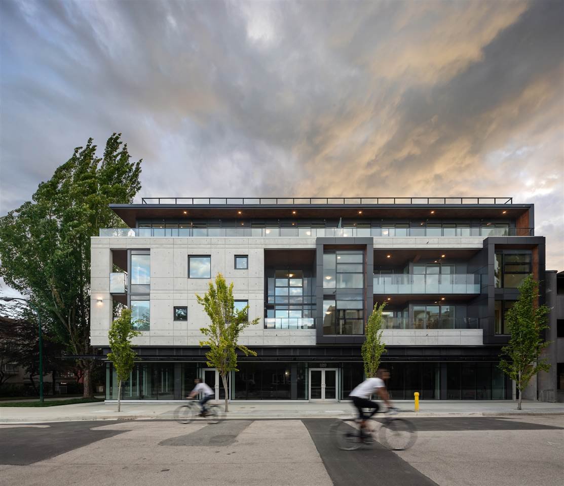 Modern apartment building with contemporary architecture, featuring large glass windows and balconies. Two people on bicycles ride past on the street in the foreground. Trees are situated in front of the building under a dramatic, cloudy sky.