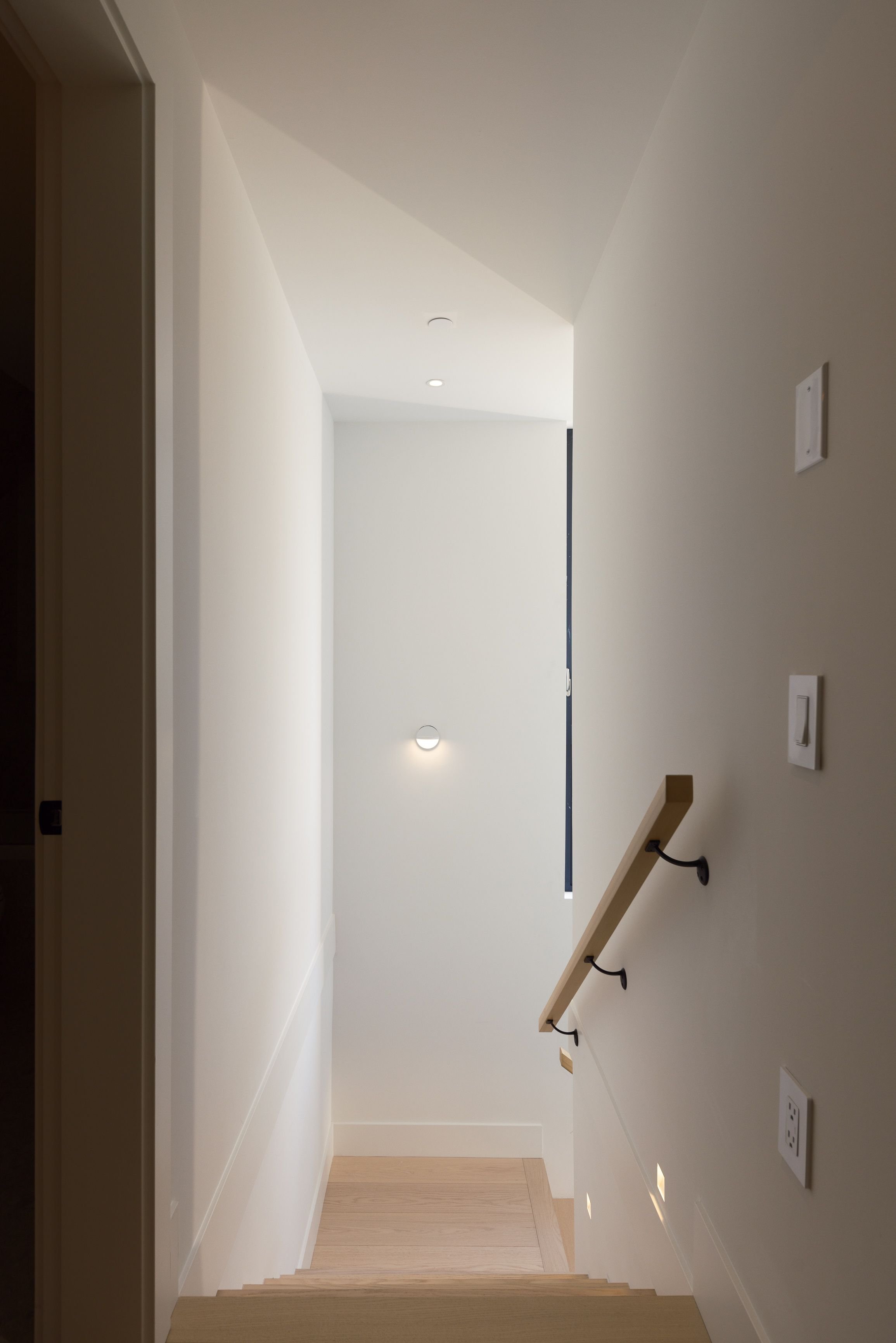 Modern staircase with wooden steps and handrail, white walls, recessed lighting, and wall-mounted light fixture.