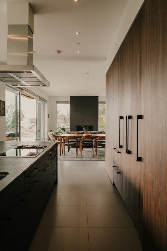 Modern kitchen interior with dark cabinetry, large windows, and a dining table in the background.