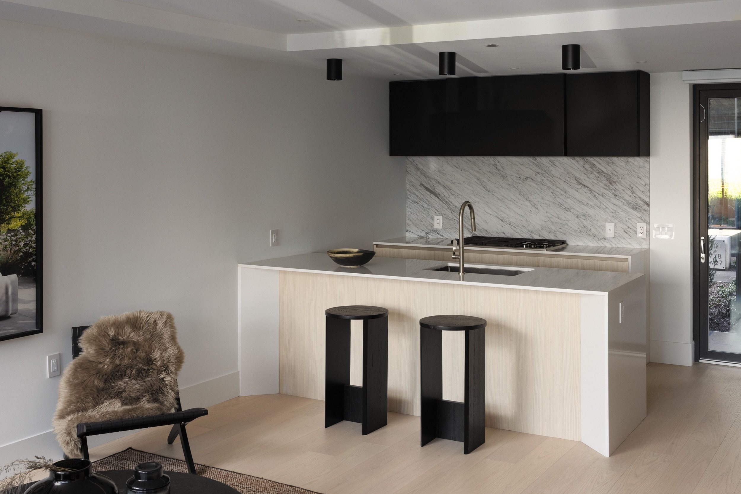 Modern kitchen interior with minimalist design, featuring a kitchen island with two black stools, a marble backsplash, black cabinets, and a stainless steel faucet. A fur-covered chair and wall art are partially visible.