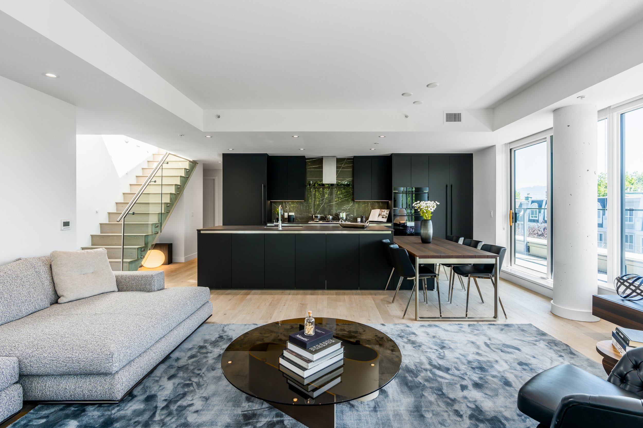 Modern open-concept living area with gray sectional sofa, black kitchen, glass staircase, wooden dining table, and large windows.