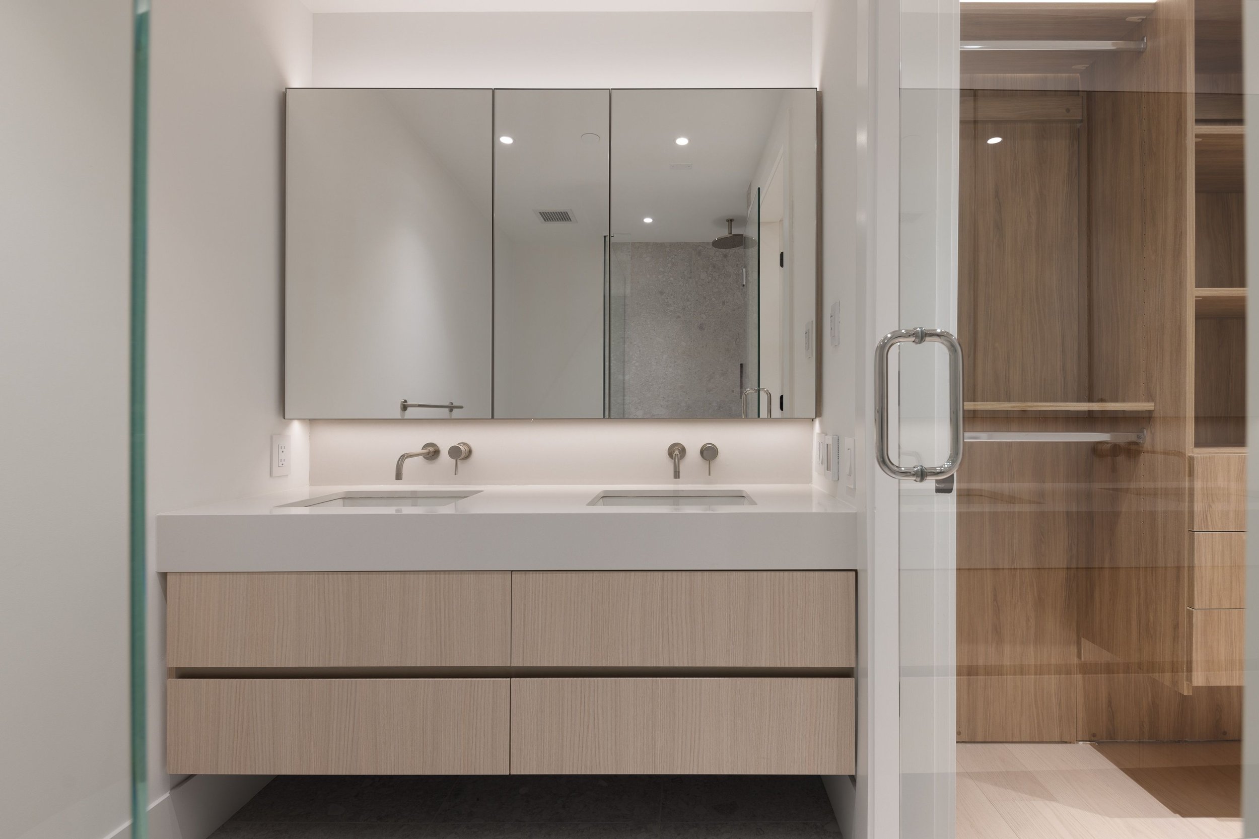 Modern bathroom with dual sinks, wall-mounted faucets, large mirrors, light wood cabinetry, and a glass door leading to a walk-in closet.