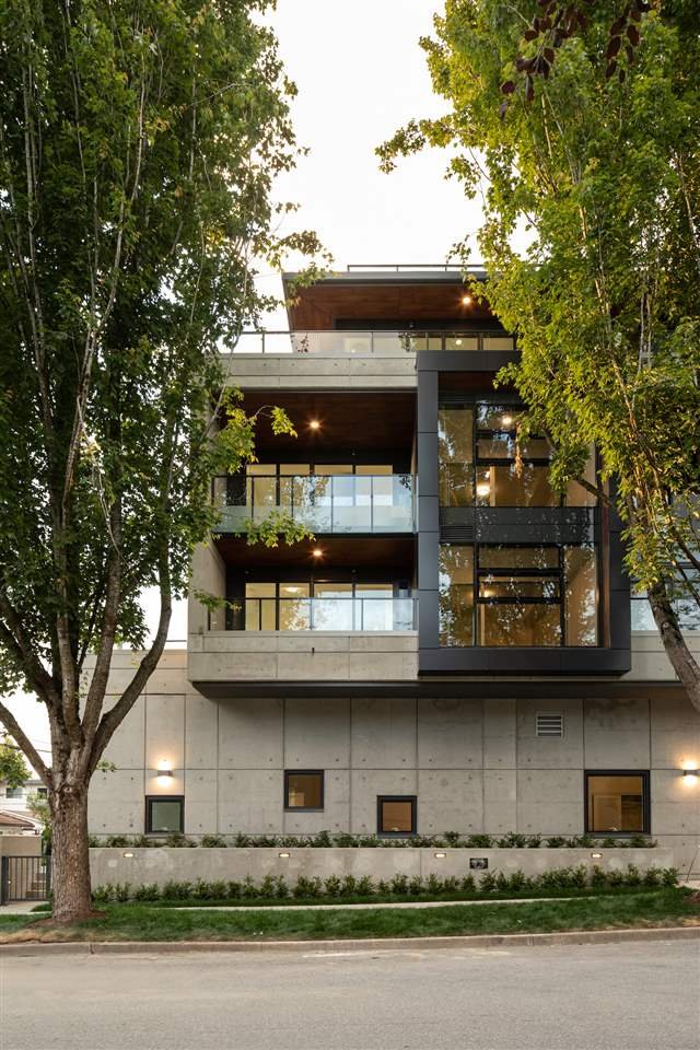 Modern multi-story building with glass facade and concrete structure, surrounded by trees and landscaping.