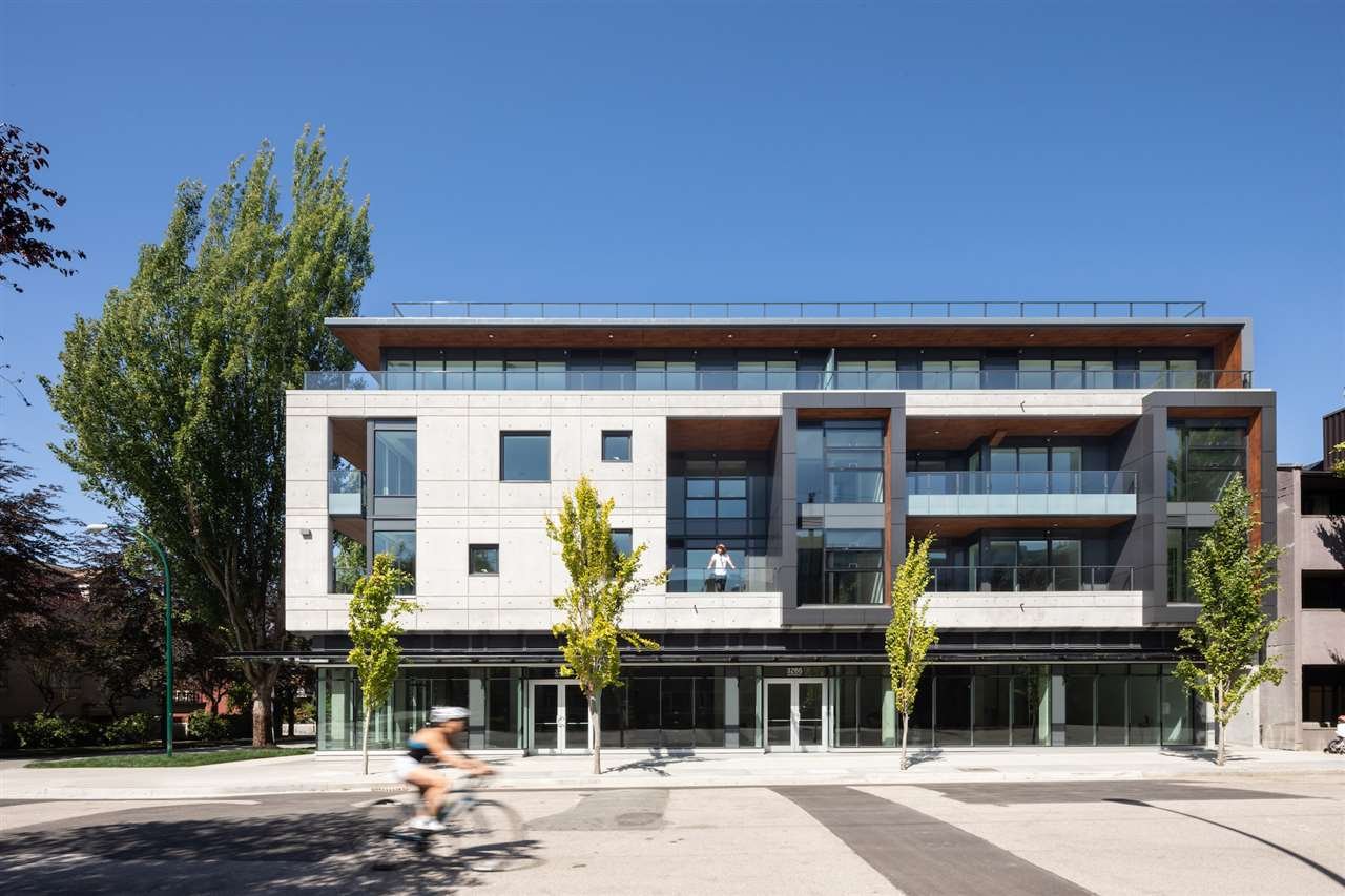 Modern multi-story building with glass and concrete facade, trees in front, a person cycling in the foreground, and a person standing on a balcony, under a clear blue sky.