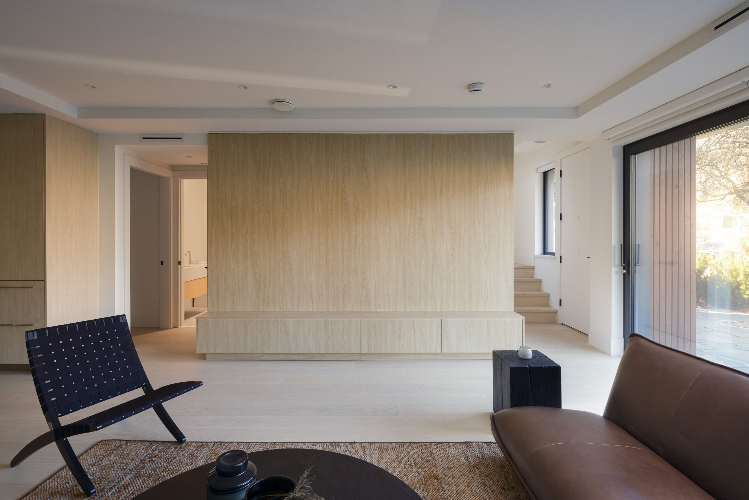 Modern minimalist living room with wooden paneling, a black woven chair, brown leather sofa, floor-to-ceiling windows, and beige flooring.