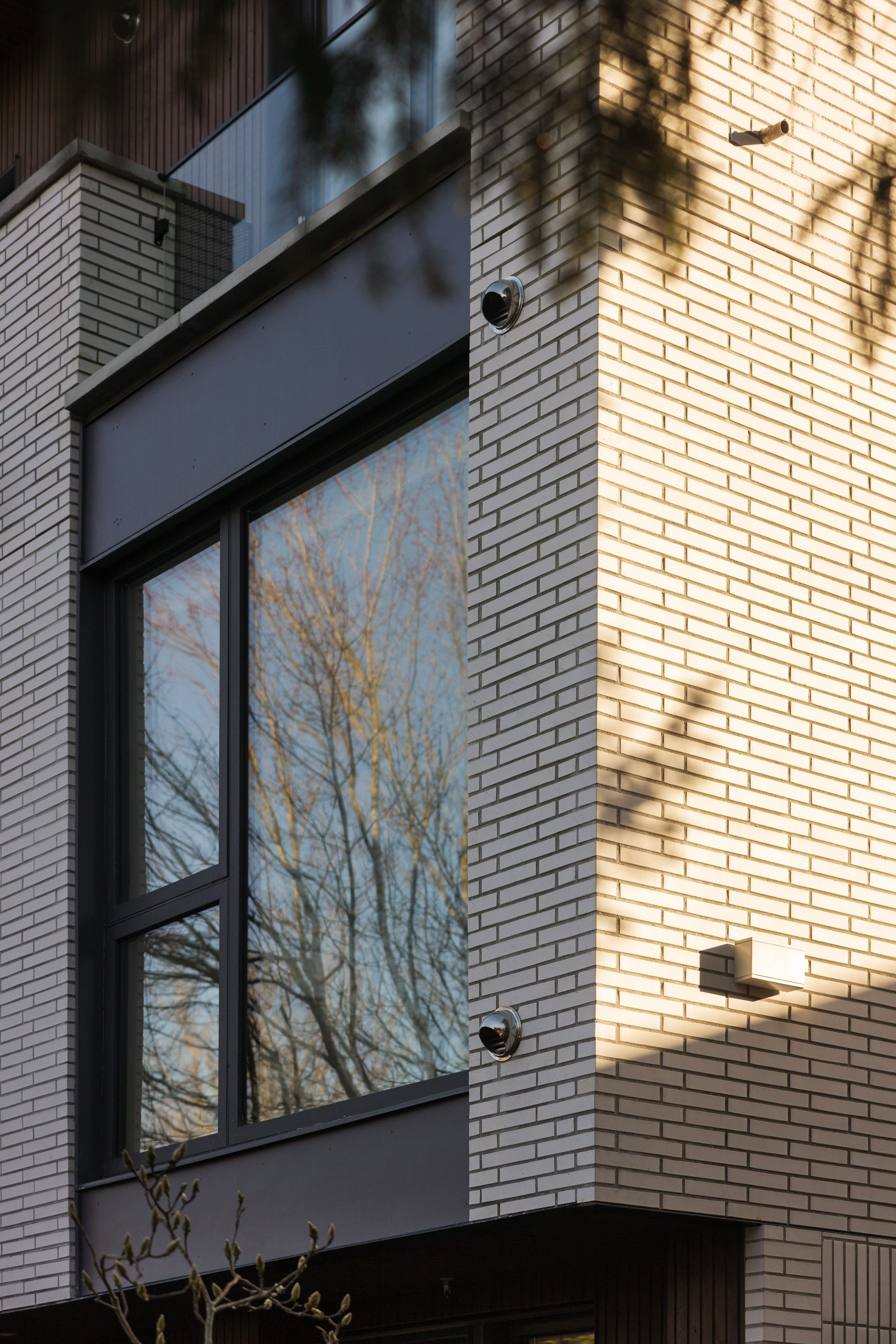 Modern brick building facade with large window reflecting bare trees, security camera, and shadows from nearby foliage.