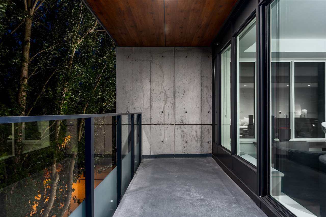 Modern apartment balcony at night with glass railing, concrete walls, and indoor view through large windows.
