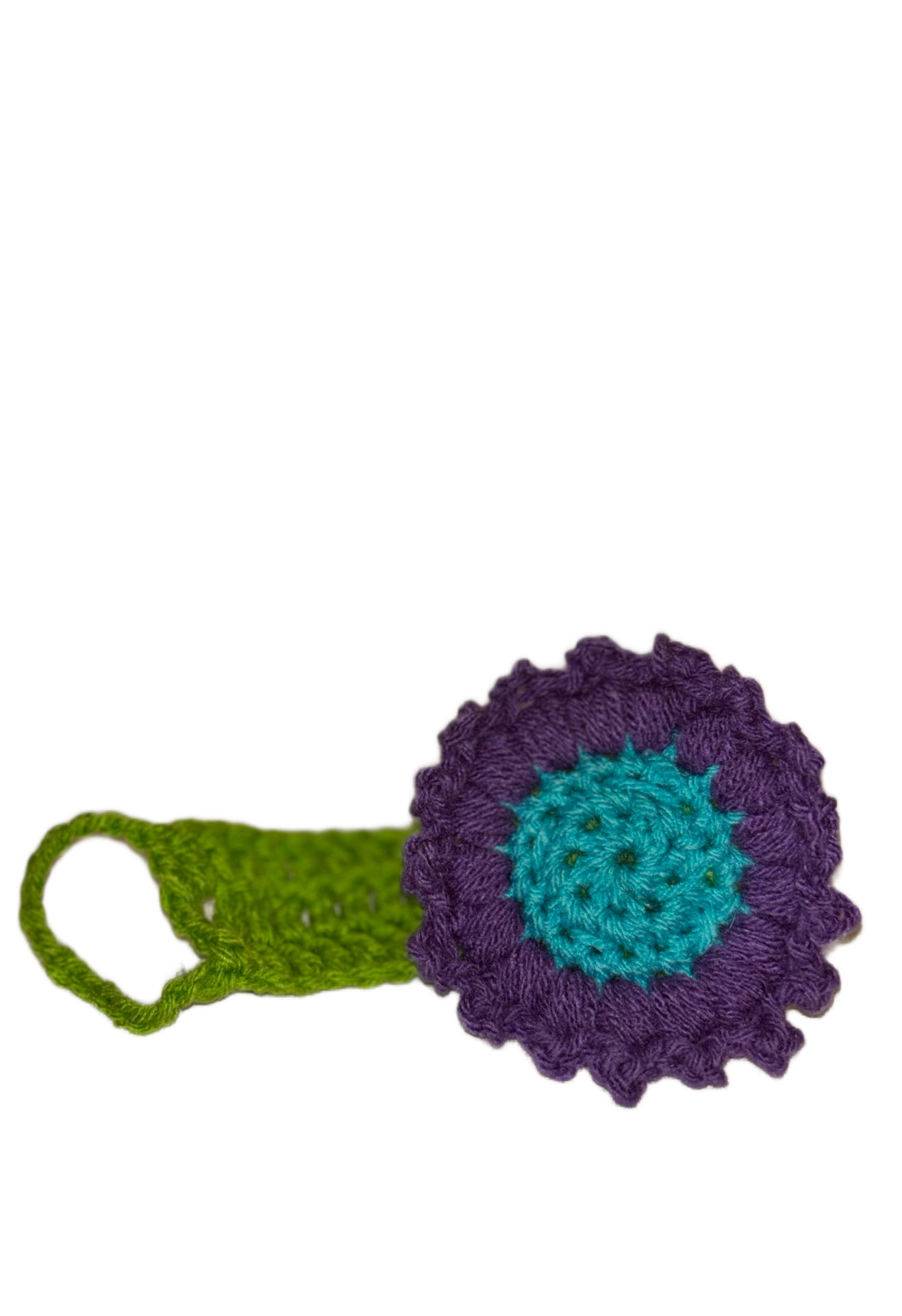 A colorful crocheted flower with a purple outer rim, a turquoise inner circle with green details, and a green stem with a loop on one side, set against a black background with white dots.