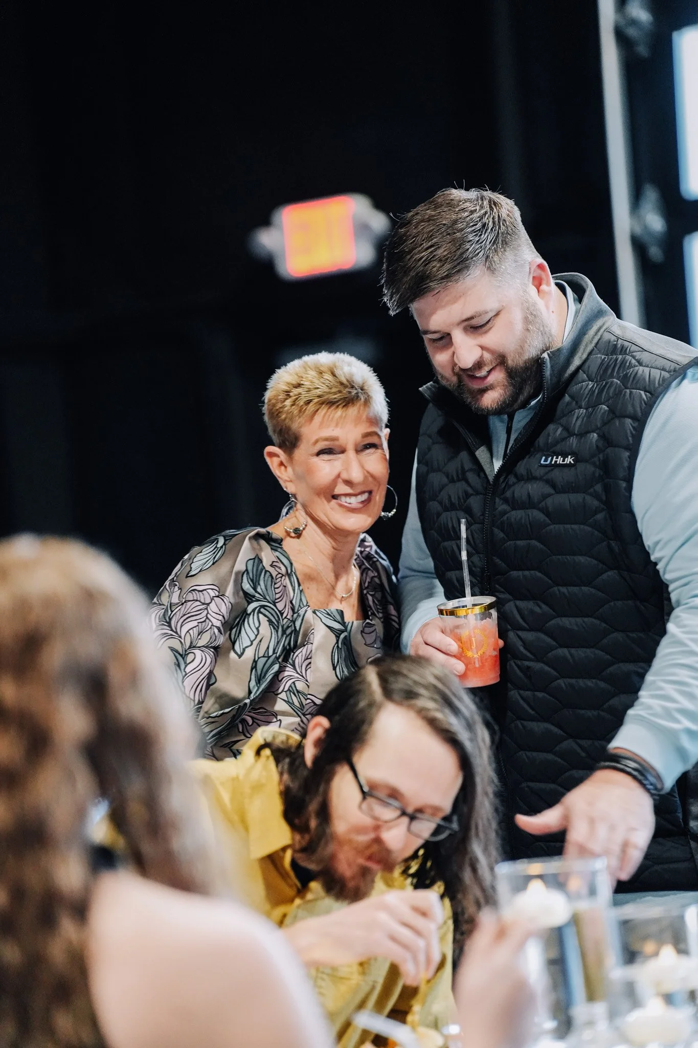 A group of people socializing at a gathering, with a woman smiling and a young man handing her a drink, and other people sitting at a table in the foreground.