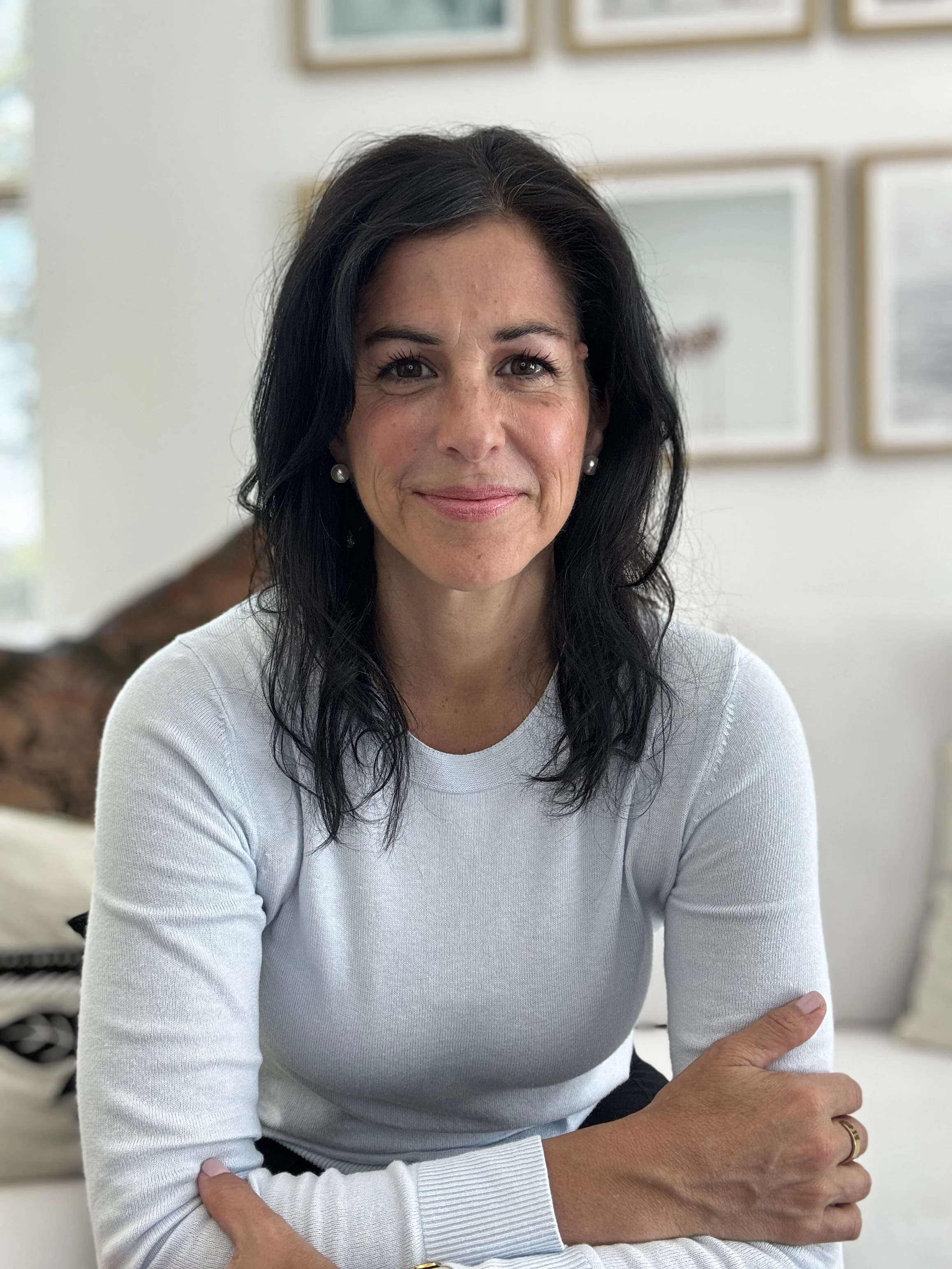 A woman with dark hair, wearing a blue sweater, sitting with her arms crossed, smiling in a well-lit room with framed artwork on the wall behind her.