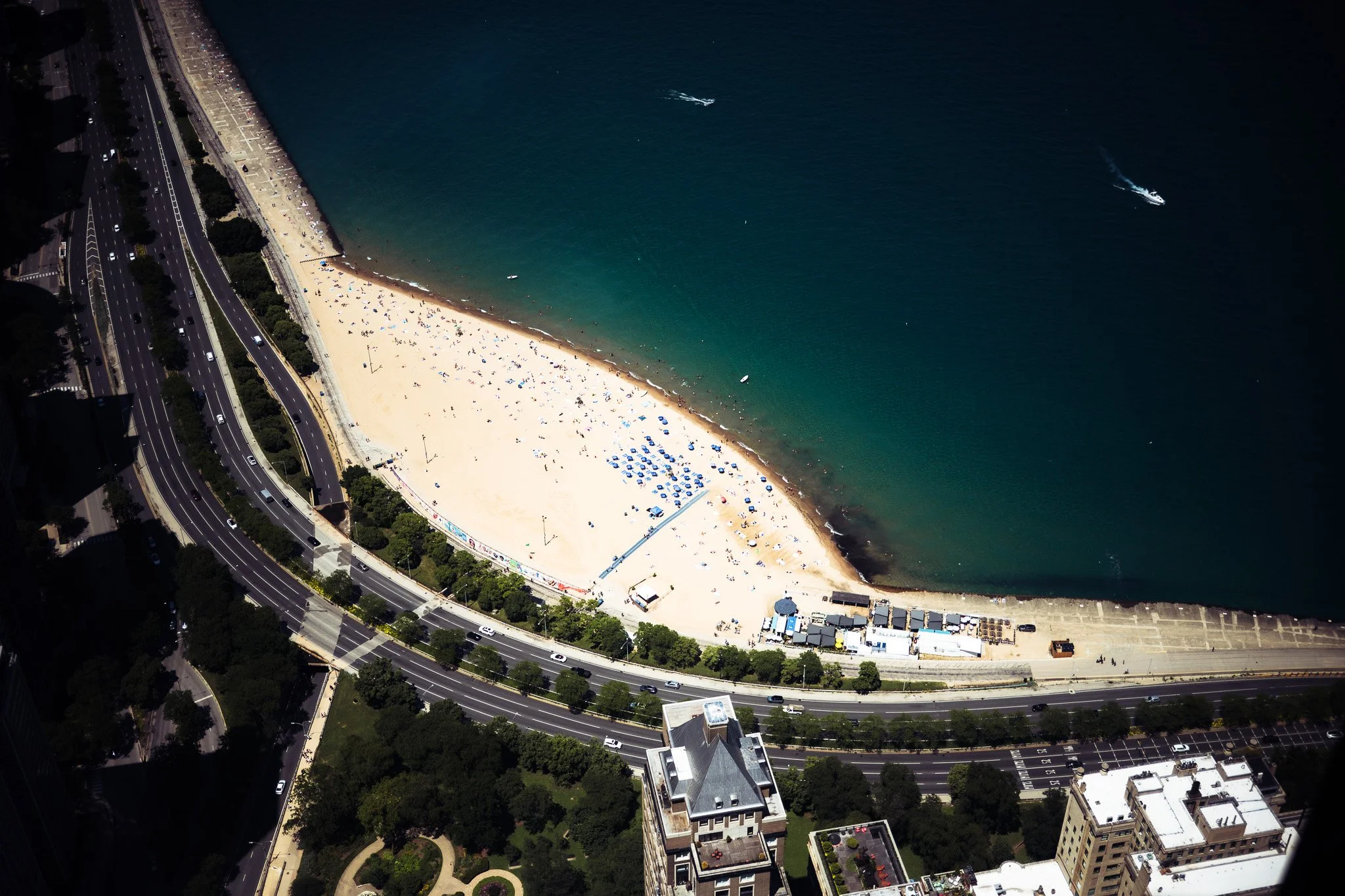 Aerial view of a crowded sandy beach beside a multi-lane coastal highway, with turquoise water, boats, and city buildings.