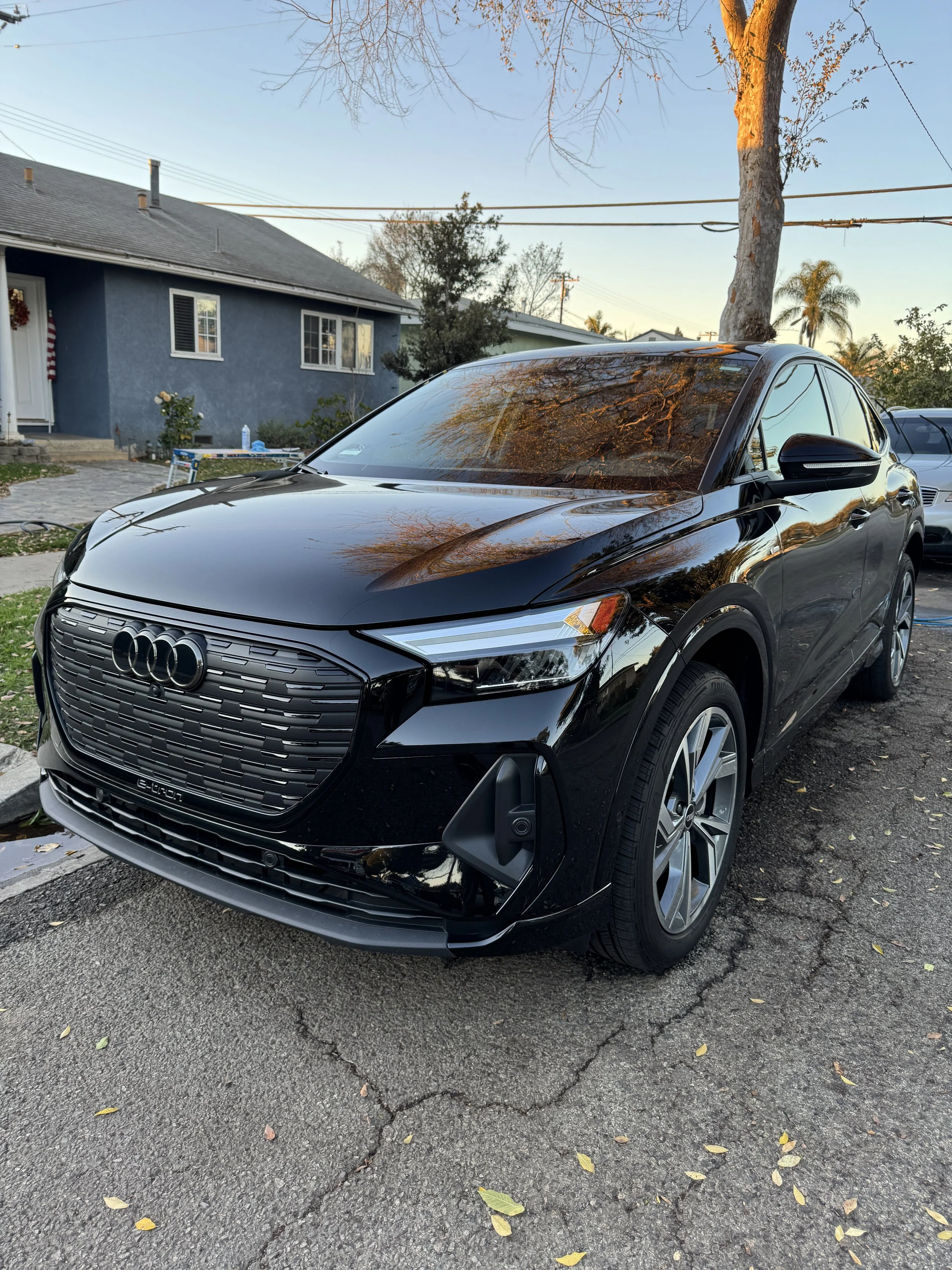 A black Audi electric vehicle parked on a cracked asphalt driveway, with a house and trees in the background during sunset.