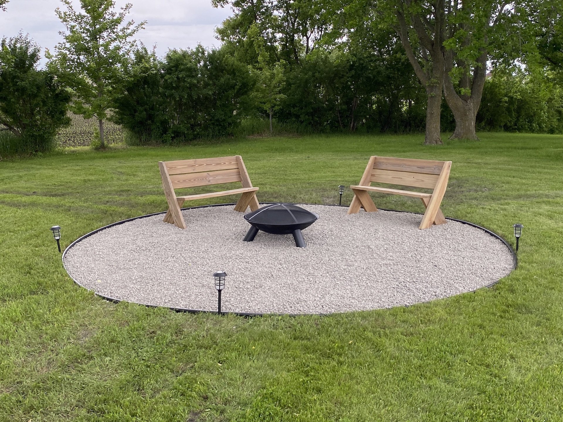 Two wooden benches facing each other with a small fire pit between them, on a gravel circle, surrounded by a grassy yard with trees in the background.