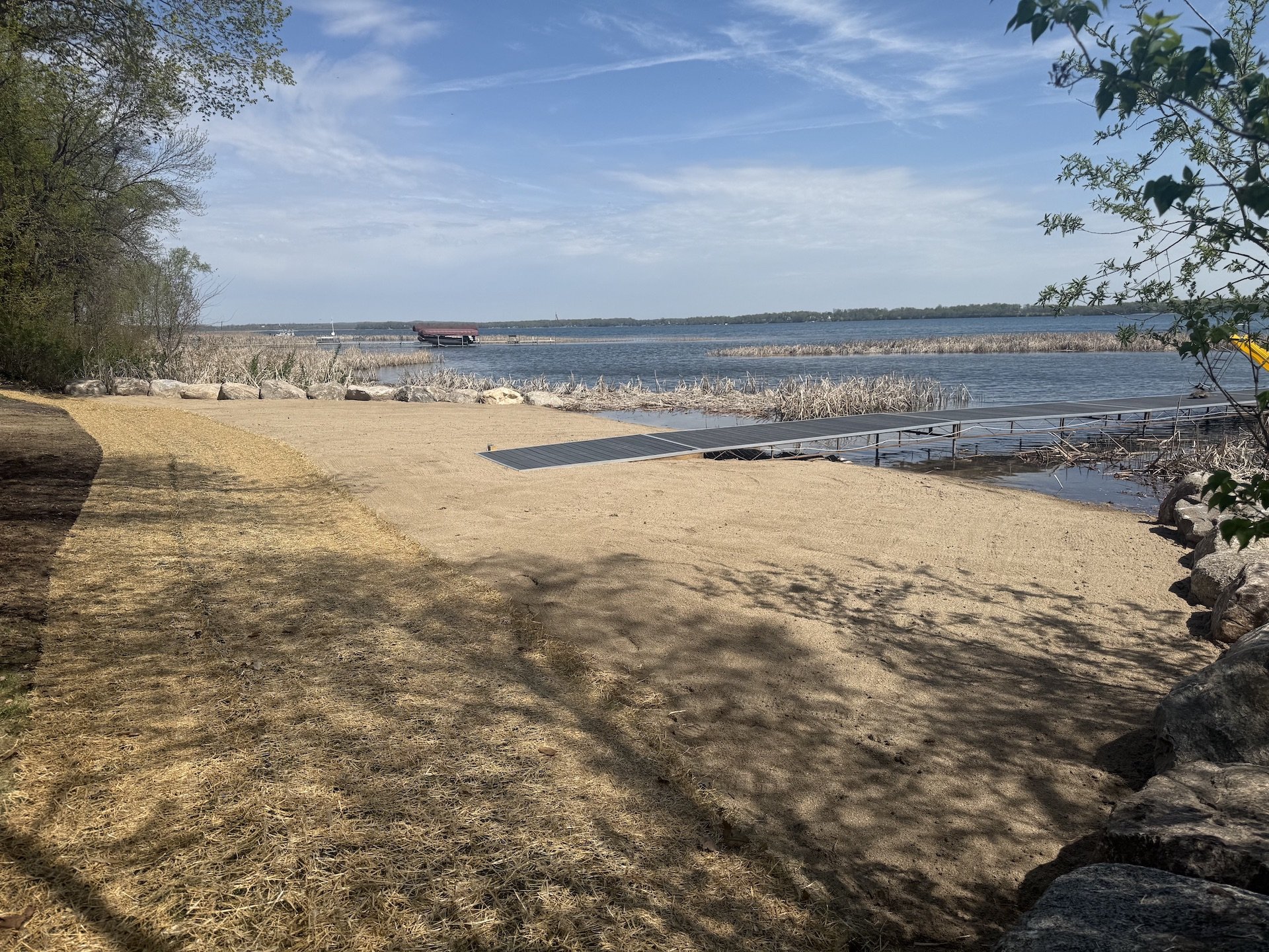 Beach with sandy shore, a metal ramp leading into the water, trees casting shadows, and a view of a lake with a dock and a boat in the distance under a partly cloudy sky.