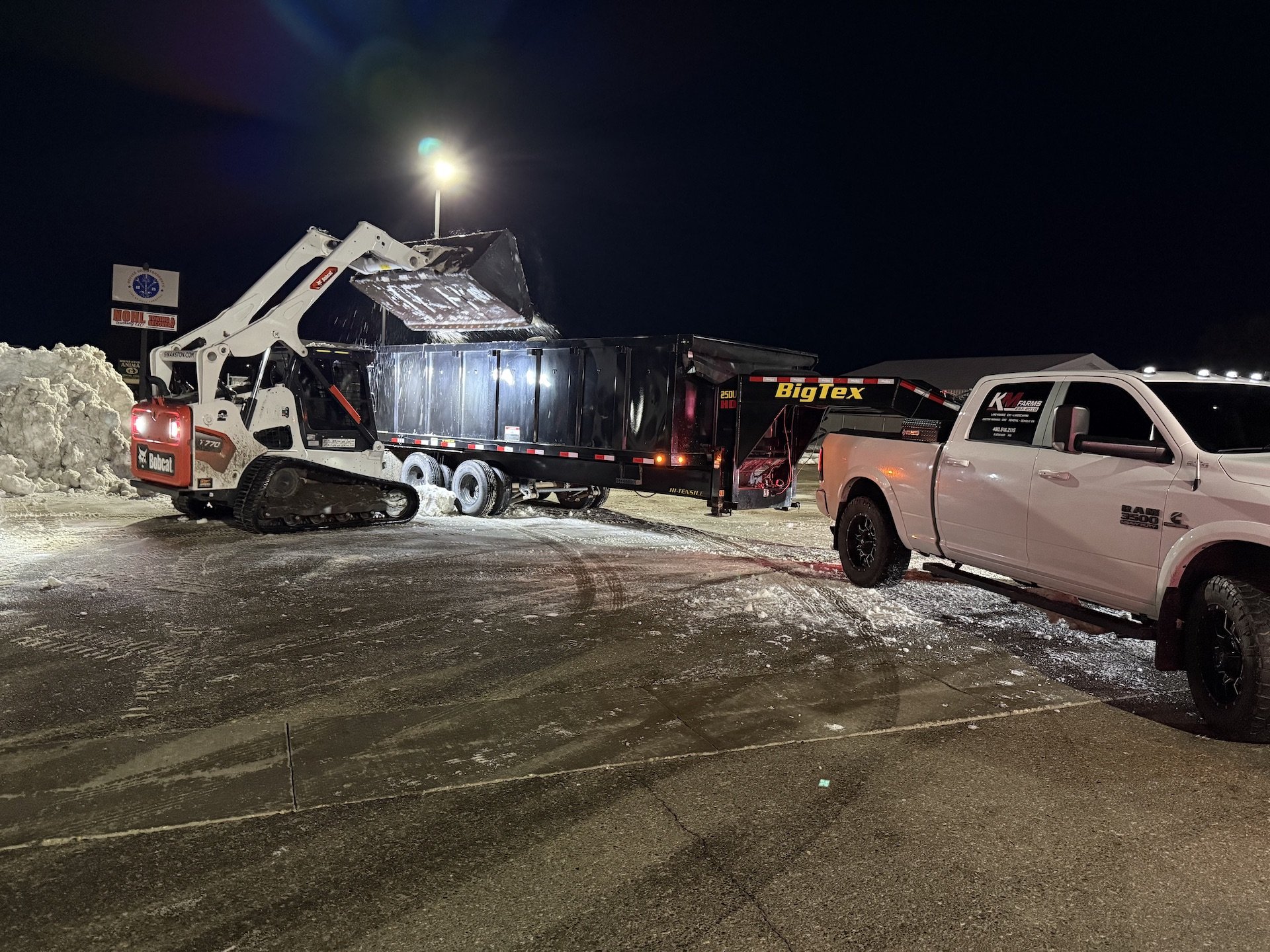 A construction vehicle loading snow into a black dump trailer attached to a white pickup truck at night