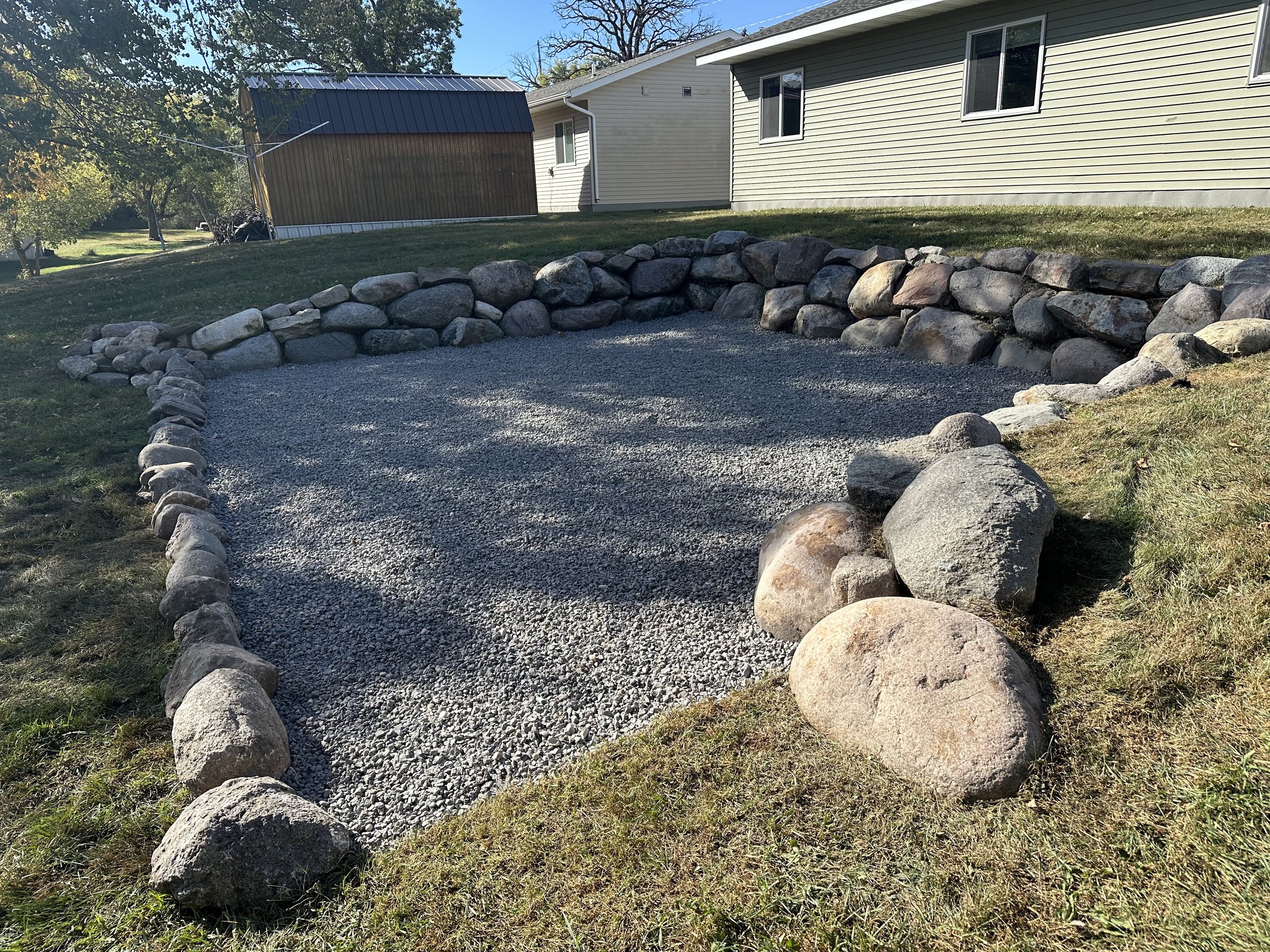 A backyard with a gravel garden bed bordered by large rocks, surrounded by grass, with houses and trees in the background under a clear sky.