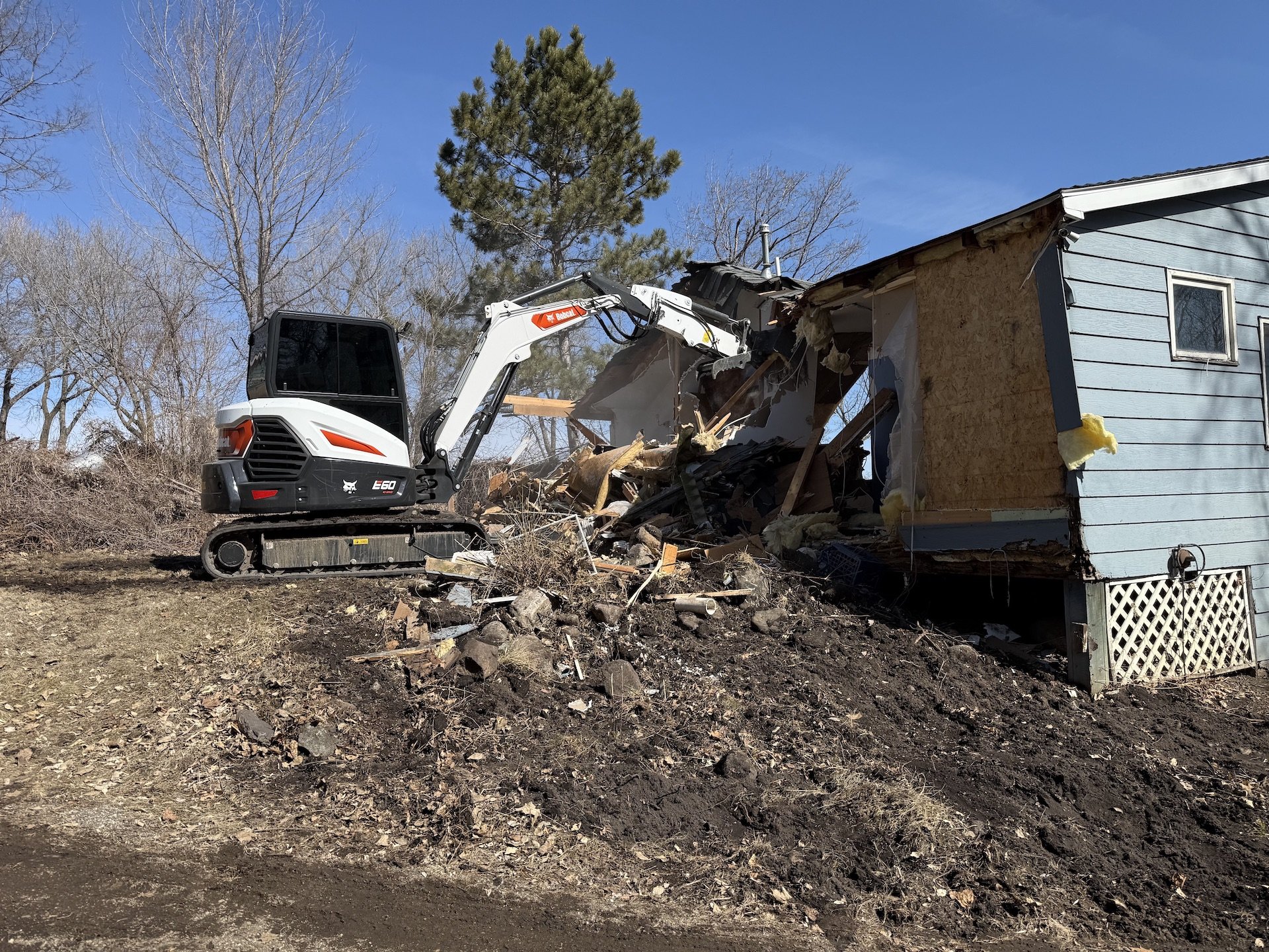 A small excavator demolishing the side of a house on a dirt lot with trees and a clear blue sky in the background.