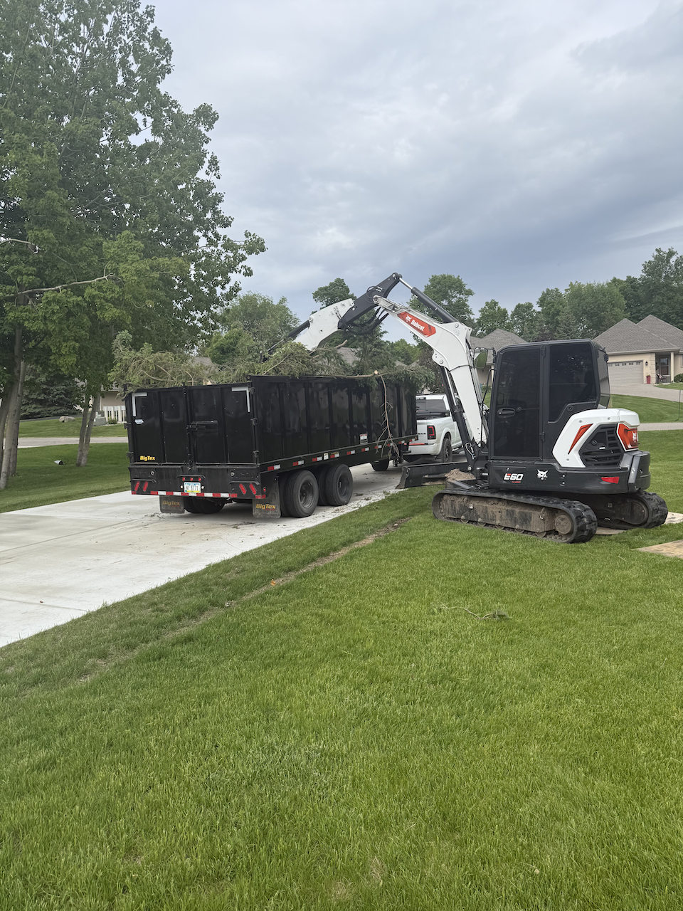 A small excavator is loading dirt or debris into a black dump trailer on a residential driveway. Green grass, trees, and houses are visible in the background under an overcast sky.