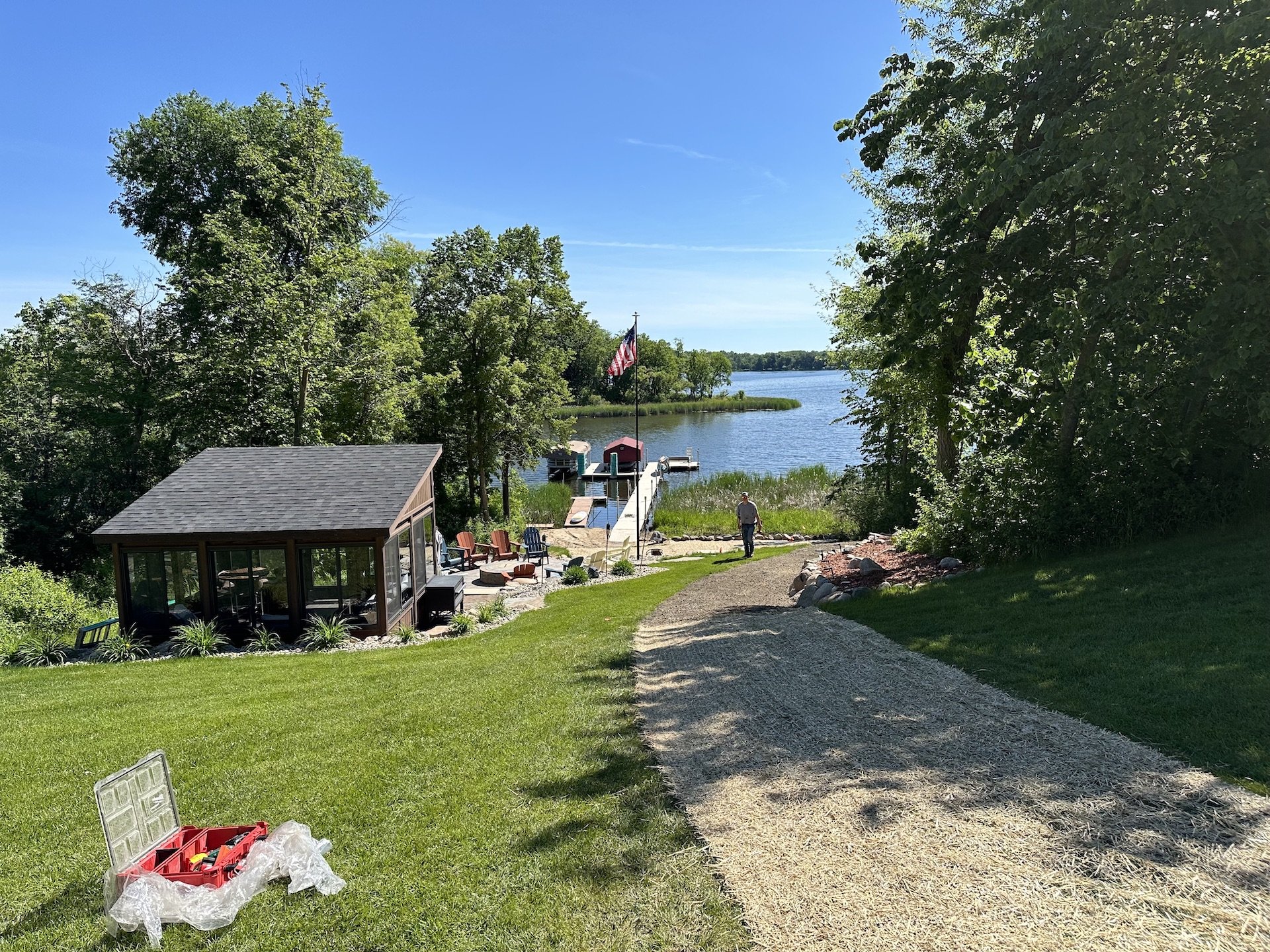 A lakeside backyard with a gravel path leading to a boat dock. There is a glass-enclosed structure, several Adirondack chairs, a person walking near the water, and trees lining the scene on a sunny day.