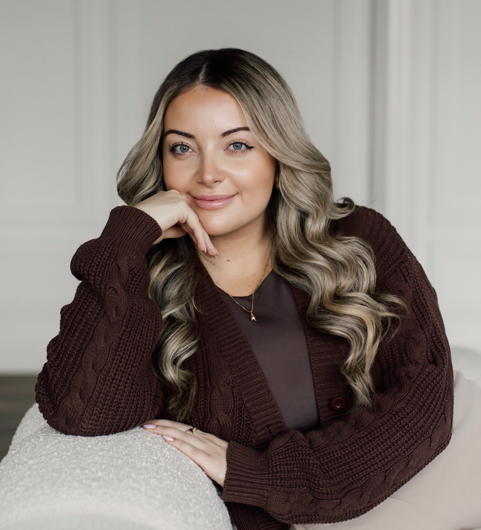 A young woman with long wavy blonde hair, wearing a dark brown cable-knit cardigan, is resting her chin on her hand and smiling at the camera in a cozy indoor setting.
