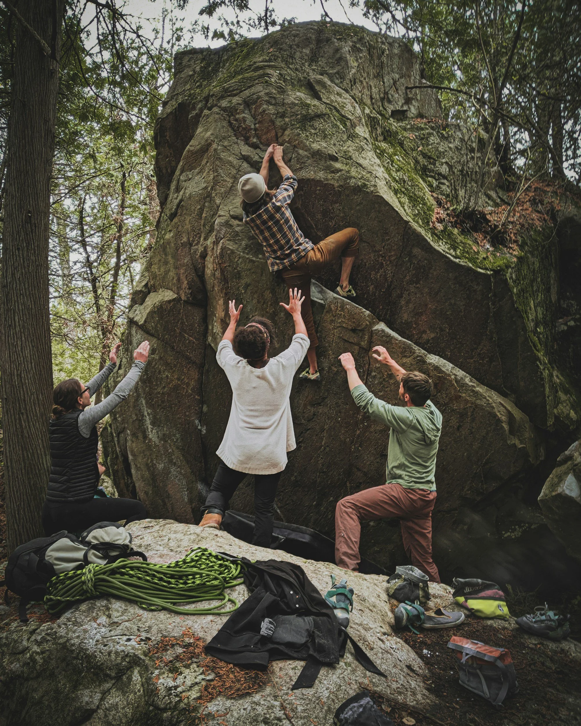 A group of four people assisting a climber on a boulder in a forested area, with climbing gear like shoes, harnesses, and ropes on the ground.