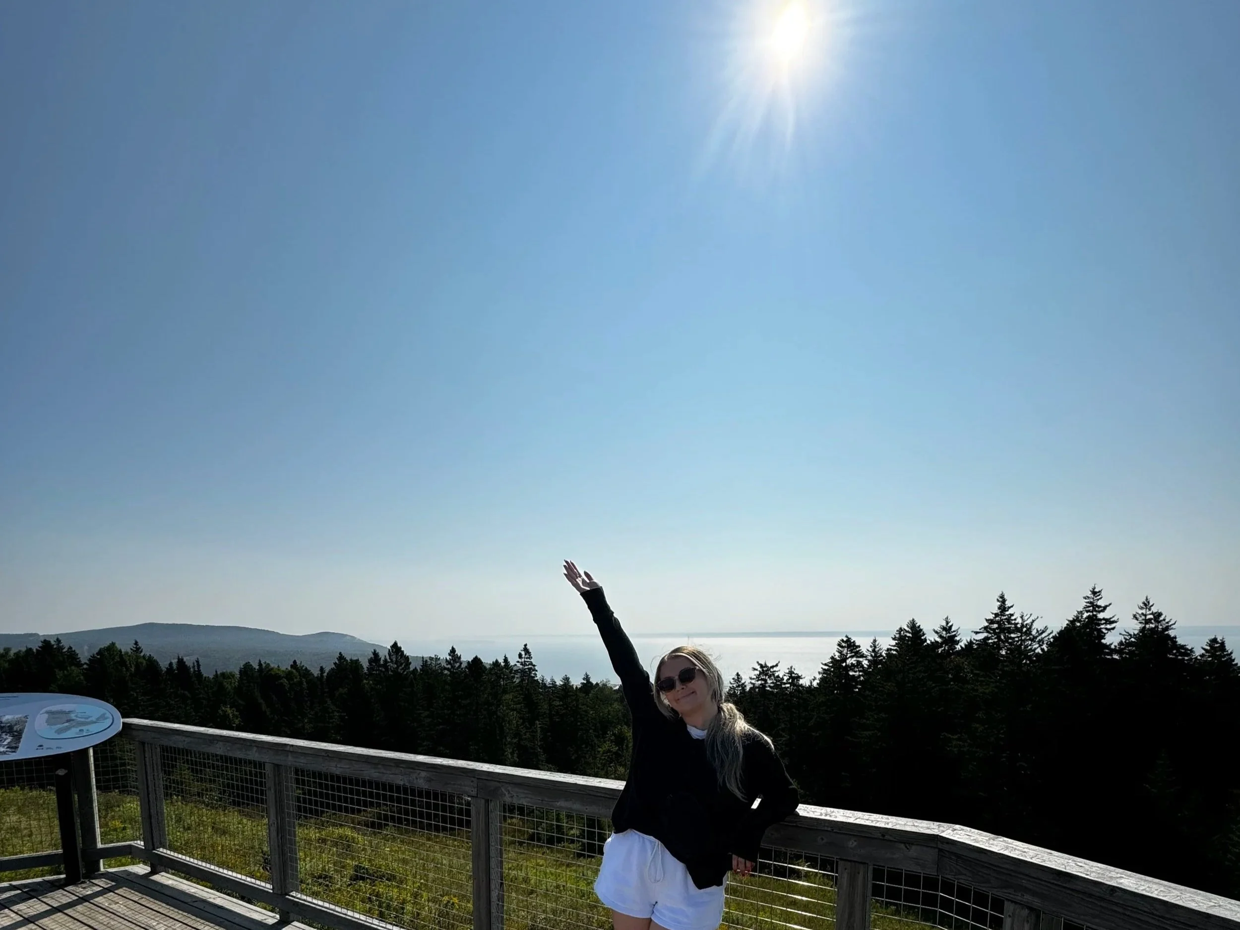 A woman with long blonde hair, wearing sunglasses, a black jacket, and white shorts, stands on a wooden viewing platform with metal railings, raising one arm toward the bright sun in a clear blue sky. Behind her are dense green trees and distant hills.