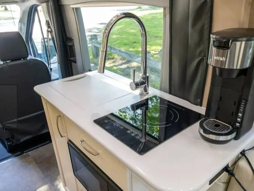 Kitchen area in camper van with a white countertop, black induction stove, stainless steel sink with high arch faucet, and a coffee maker on the right. Window shows green grass outside.