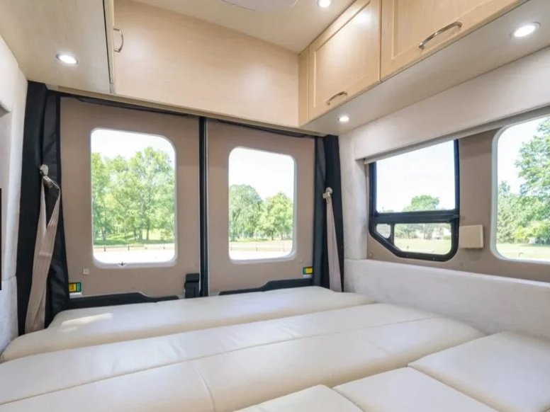 Interior of a camper van with large windows, a plush white bed, and wooden cabinets on the ceiling.