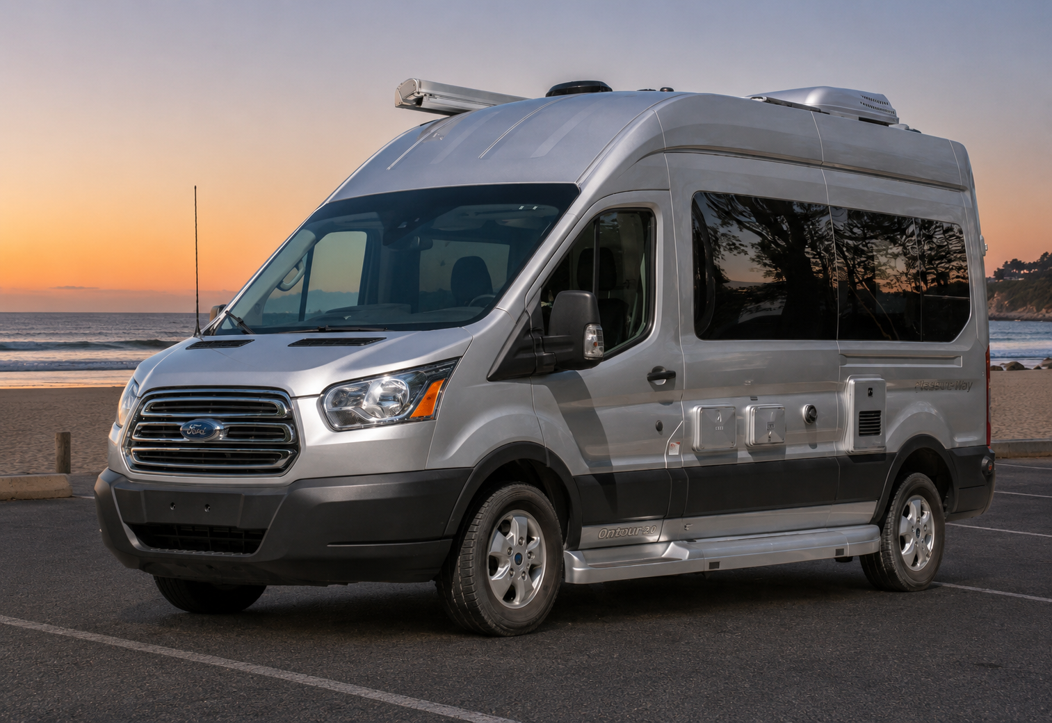 A silver Ford Transit van parked on a beach parking lot during sunset, with the ocean and a distant shoreline in the background.