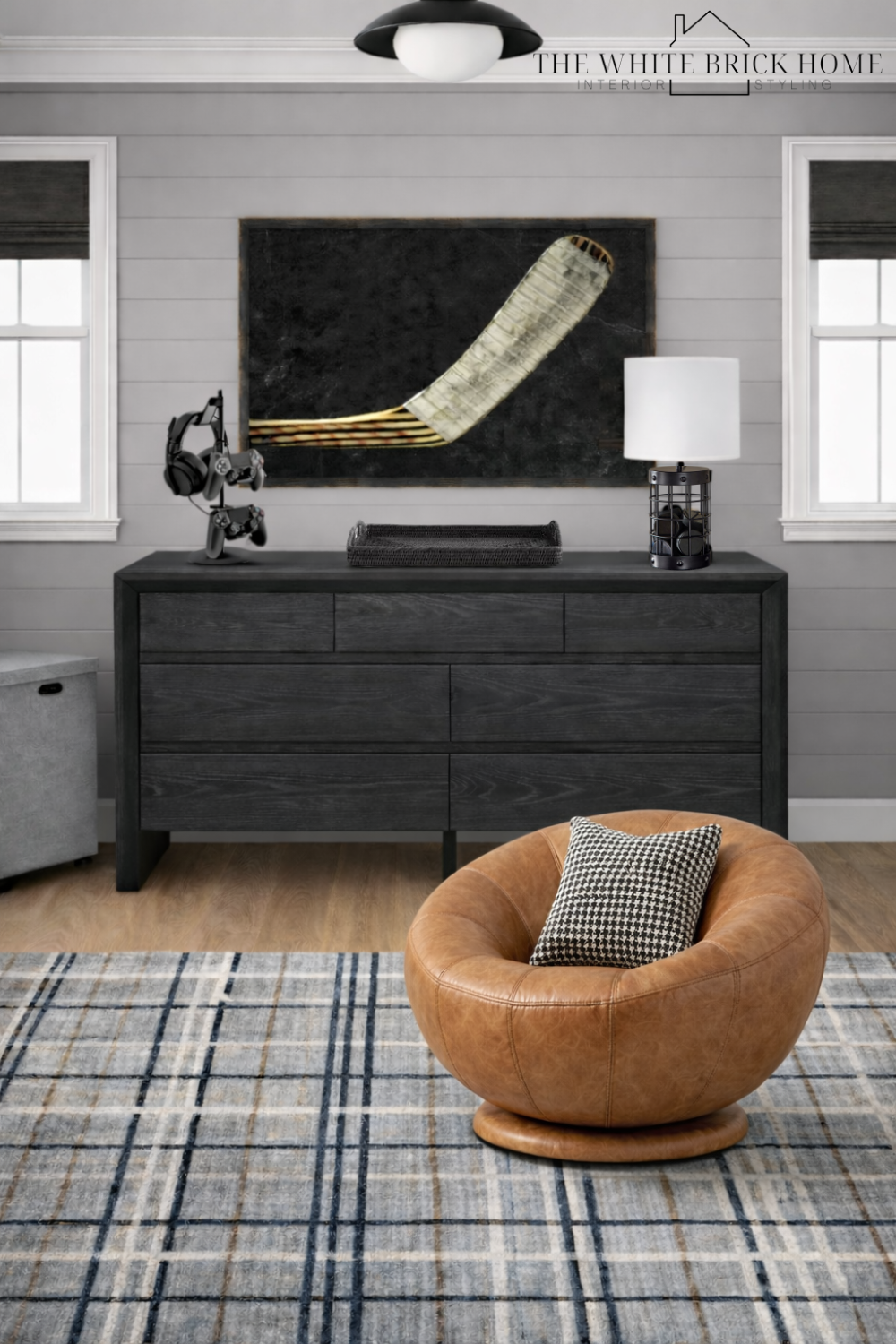 Modern interior room with gray walls, black dresser, large blackboard with a curled wooden object, white table lamp, black headphone stand, a houndstooth pillow on a round tan leather chair, plaid rug, and two windows.