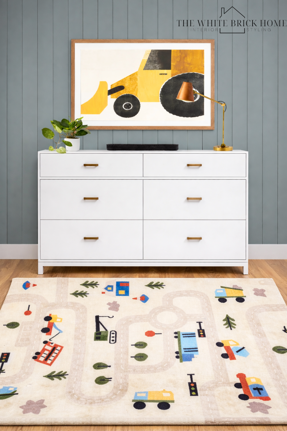 A white dresser with gold handles against a gray wall with vertical paneling, decorated with a potted green plant and a gold desk lamp. Above, a framed abstract painting of a yellow construction vehicle. The floor features a colorful children's rug w