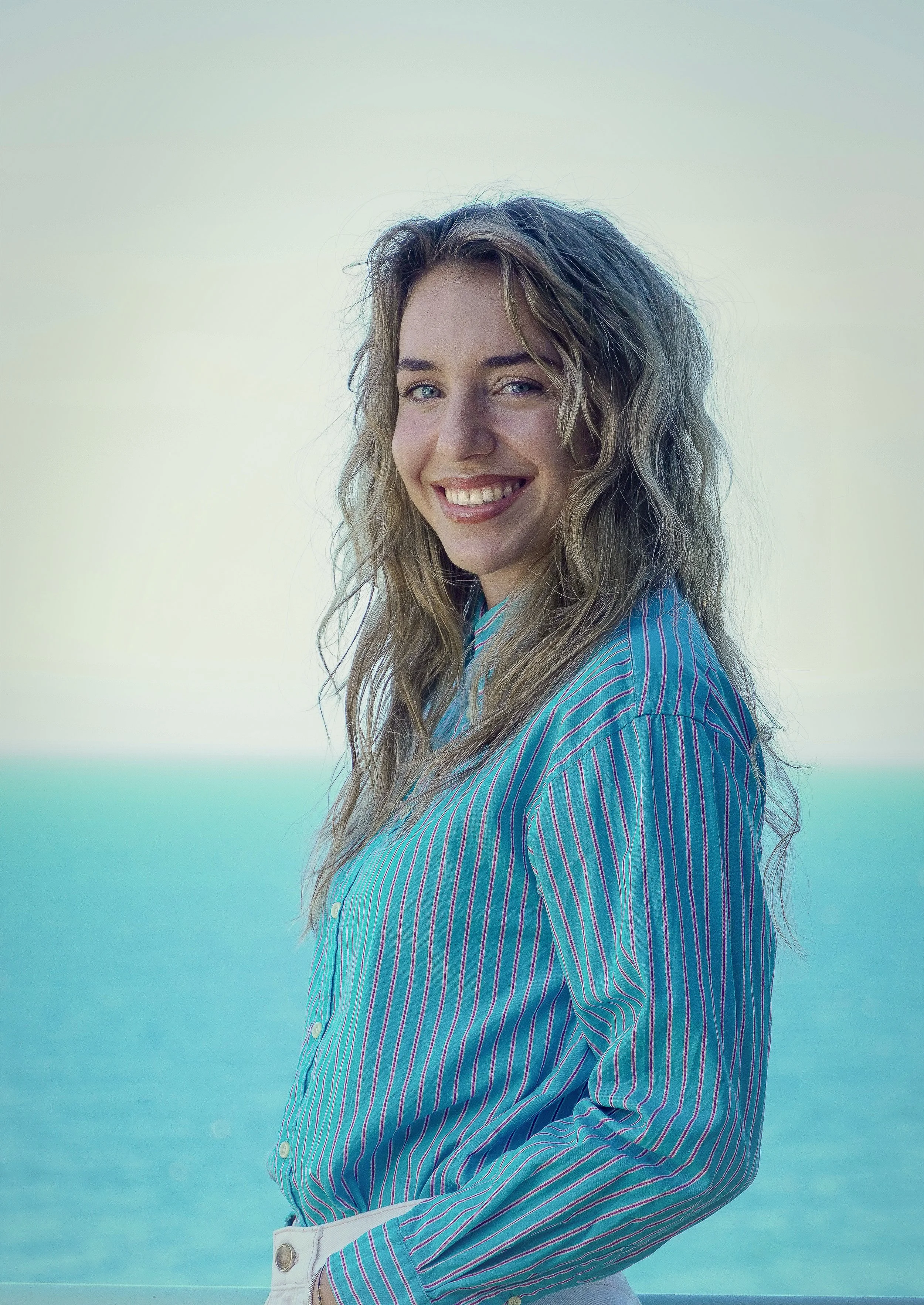 Smiling person with long hair wearing a blue striped shirt, standing in front of a bright blue ocean background.