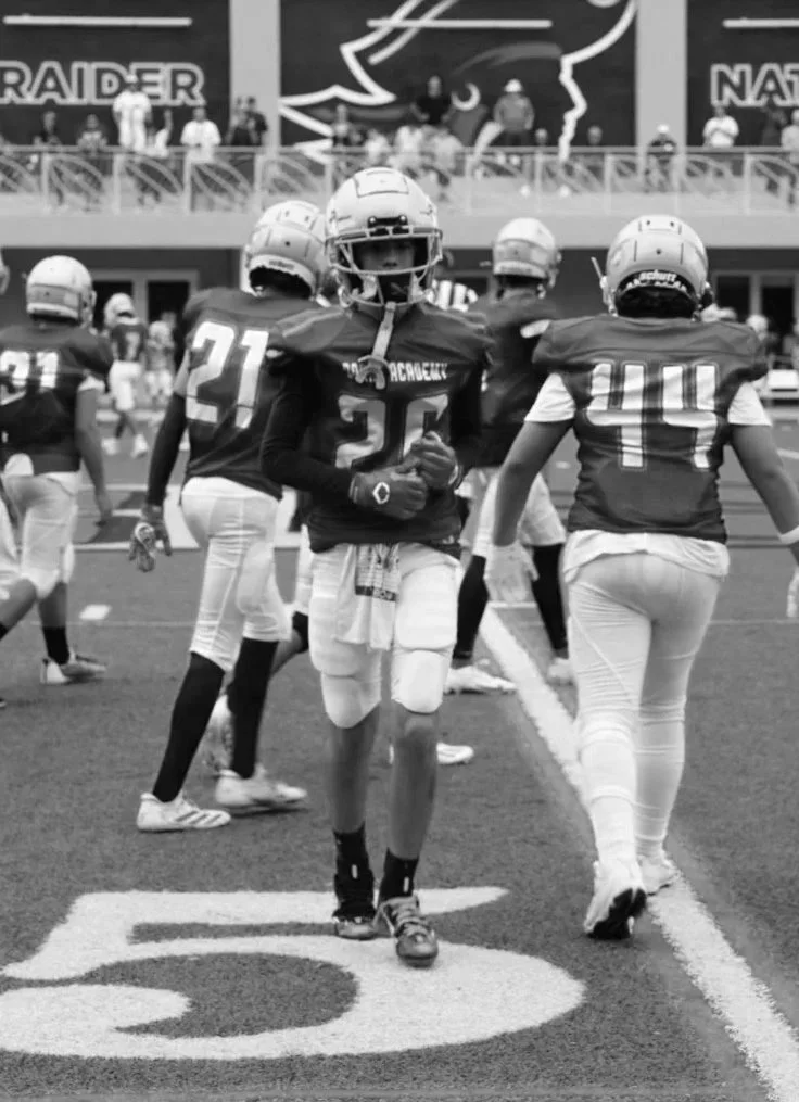 Football players on the field during a game, with one player in focus and others in background, stadium with spectators and team logo visible.