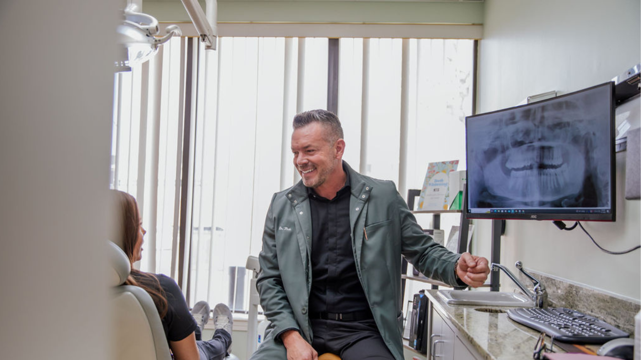A male dentist smiling and talking to a female patient seated in a dental chair during a dental appointment in an office with vertical blinds and dental equipment.