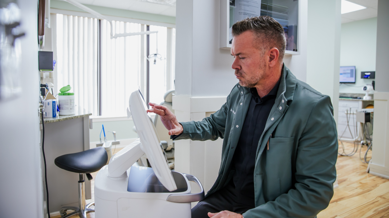 A man in a dark jacket sitting in a medical room, looking at a medical device or scanner.