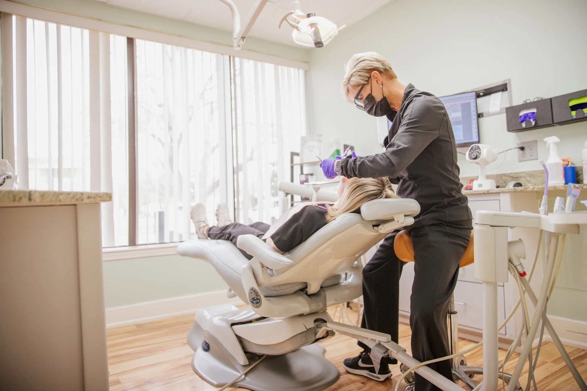 A dentist or dental hygenist works on a patient lying in a dental chair in a well-lit operatory.