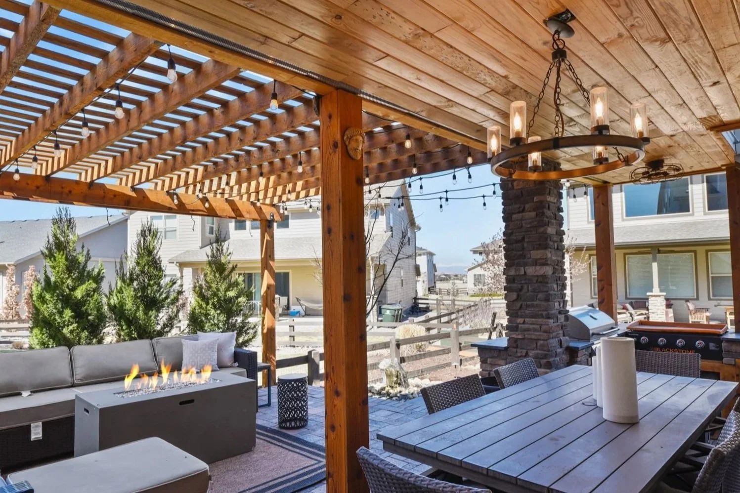 Outdoor kitchen area with a granite counter, small sink, and a stainless steel grill, surrounded by stacked stone and wooden cabinet doors, with trees and a grassy backyard in the background.