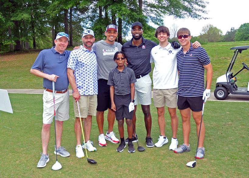 A group of seven people standing together on a golf course. They are dressed in casual golf attire, holding golf clubs. The setting includes green grass and trees, with a golf cart visible in the background.