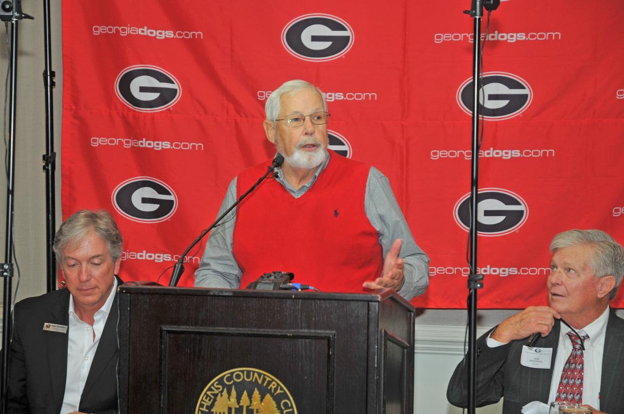 A man speaking at a podium with a University of Georgia backdrop, wearing a red vest.